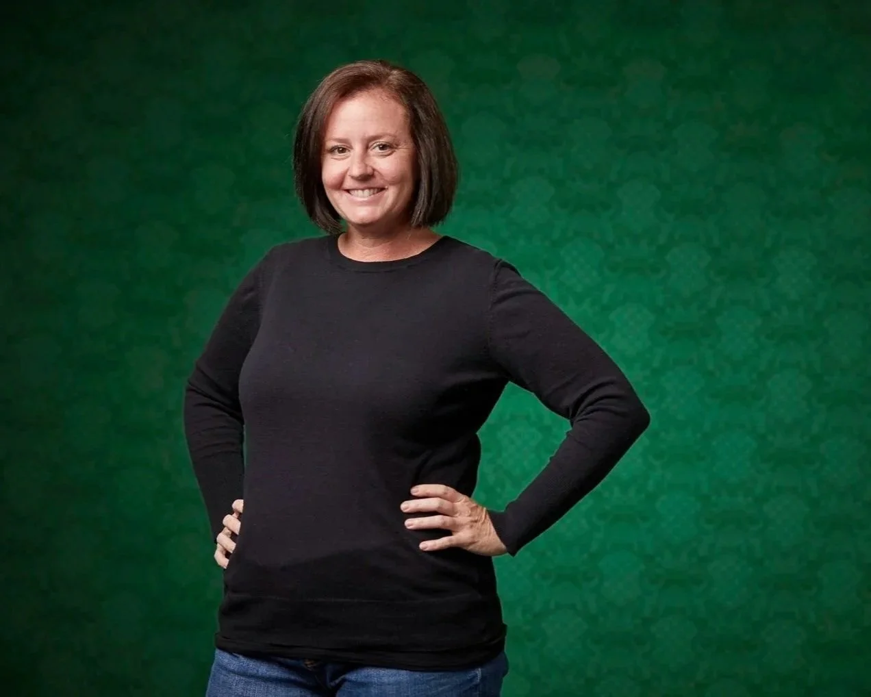 Erin Rue, certified CPA for small businesses, in a black long-sleeve shirt standing with hands on hips, smiling against a green textured background.