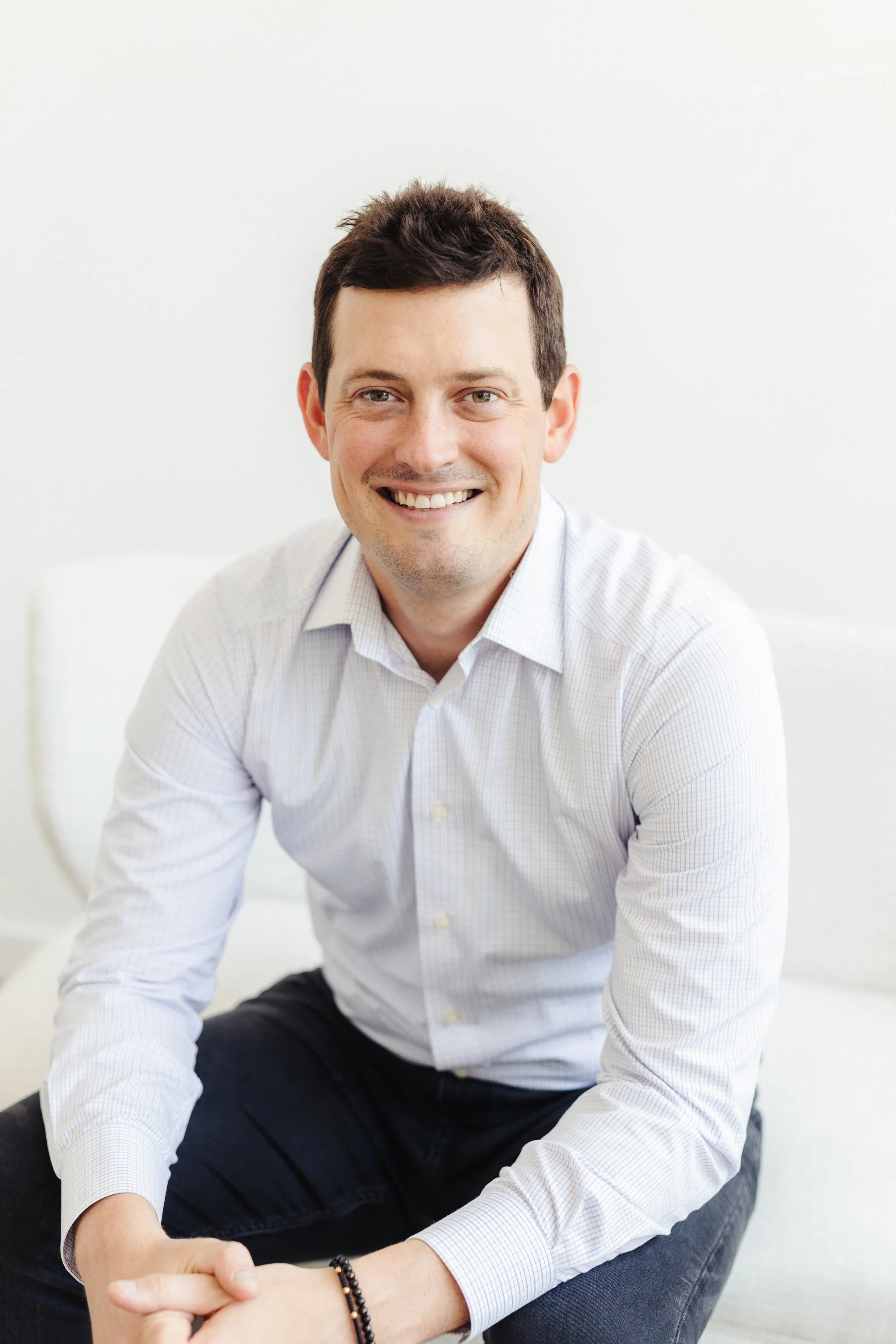 A smiling man with short dark hair wearing a light-colored button-up shirt and dark pants, sitting comfortably with one arm resting on his knee, against a plain white background.