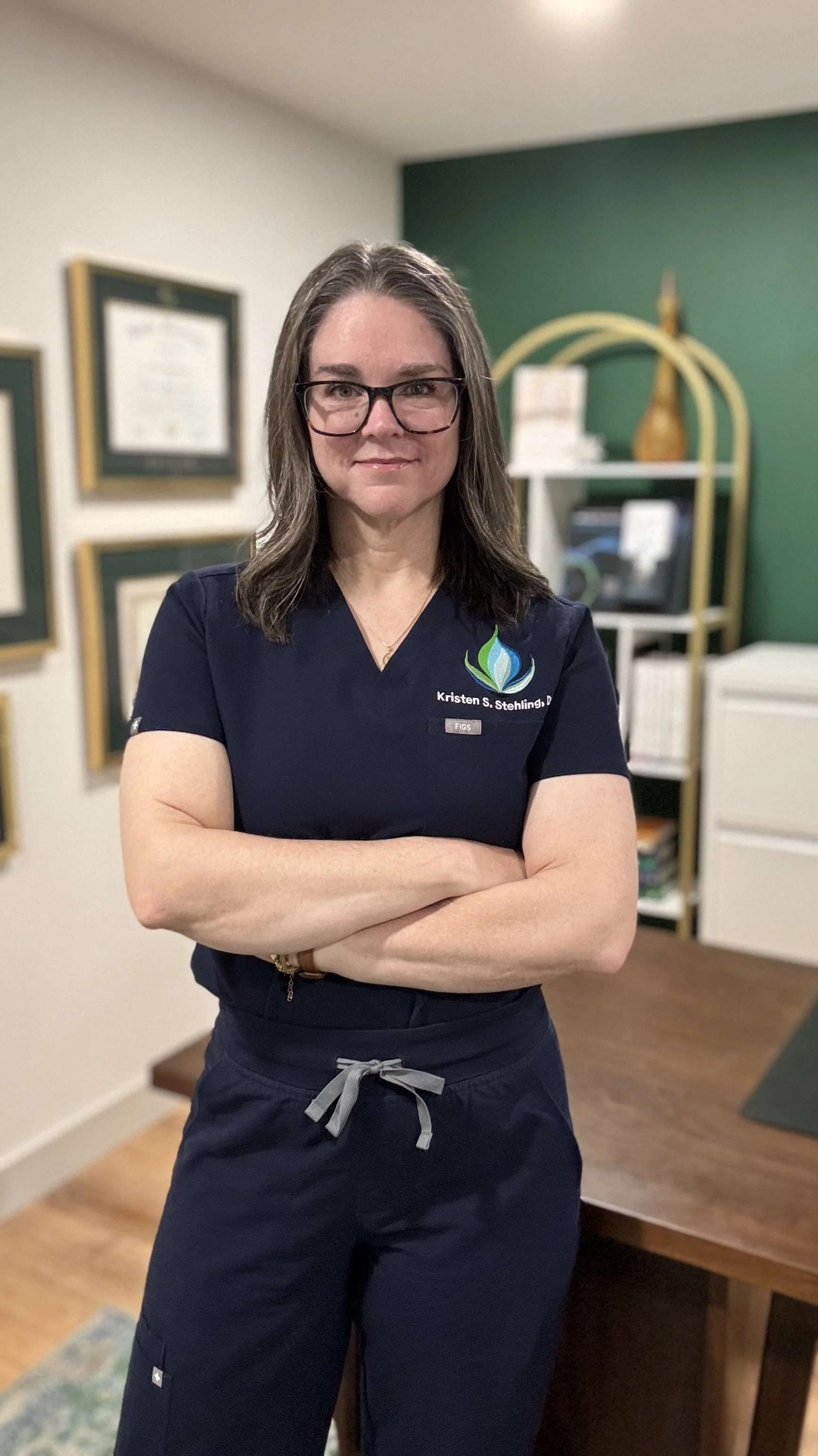 A woman with glasses and shoulder-length hair, wearing a navy medical uniform, stands with her arms crossed in an office or clinic setting. There are framed certificates on the wall and shelves with decorative items and files in the background.