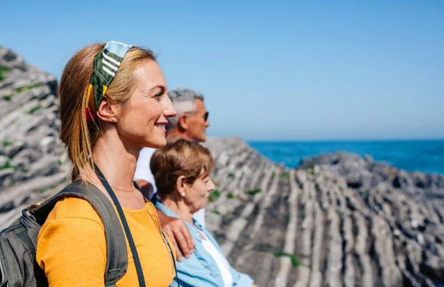 A group of four people, including a woman, an older man, an older woman, and another woman, sitting outdoors on rocky terrain near the ocean. They are enjoying the view and smiling.