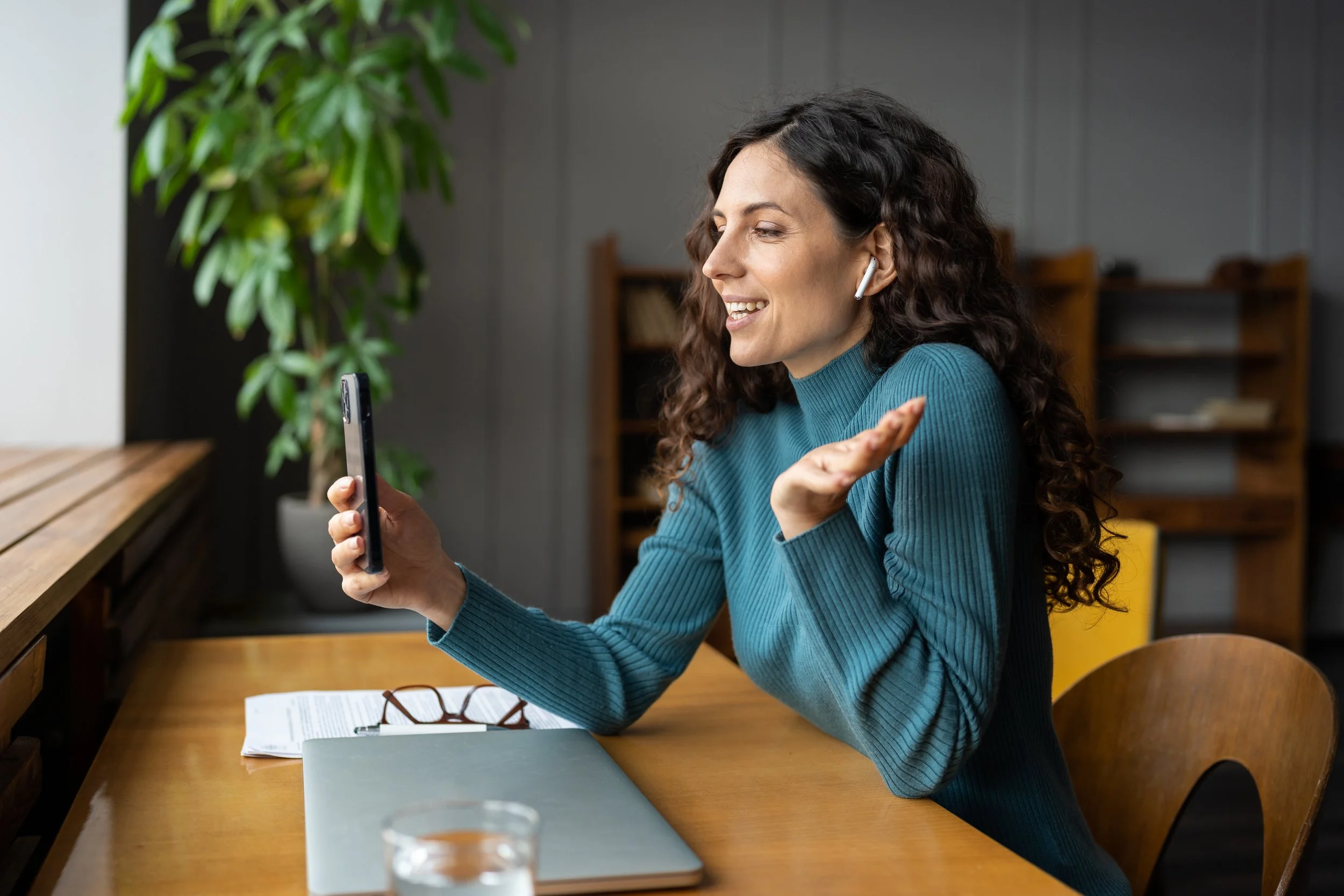 A woman sitting at a wooden desk, wearing blue sweater and wireless earbuds, smiling and gesturing while talking to someone on her phone.
