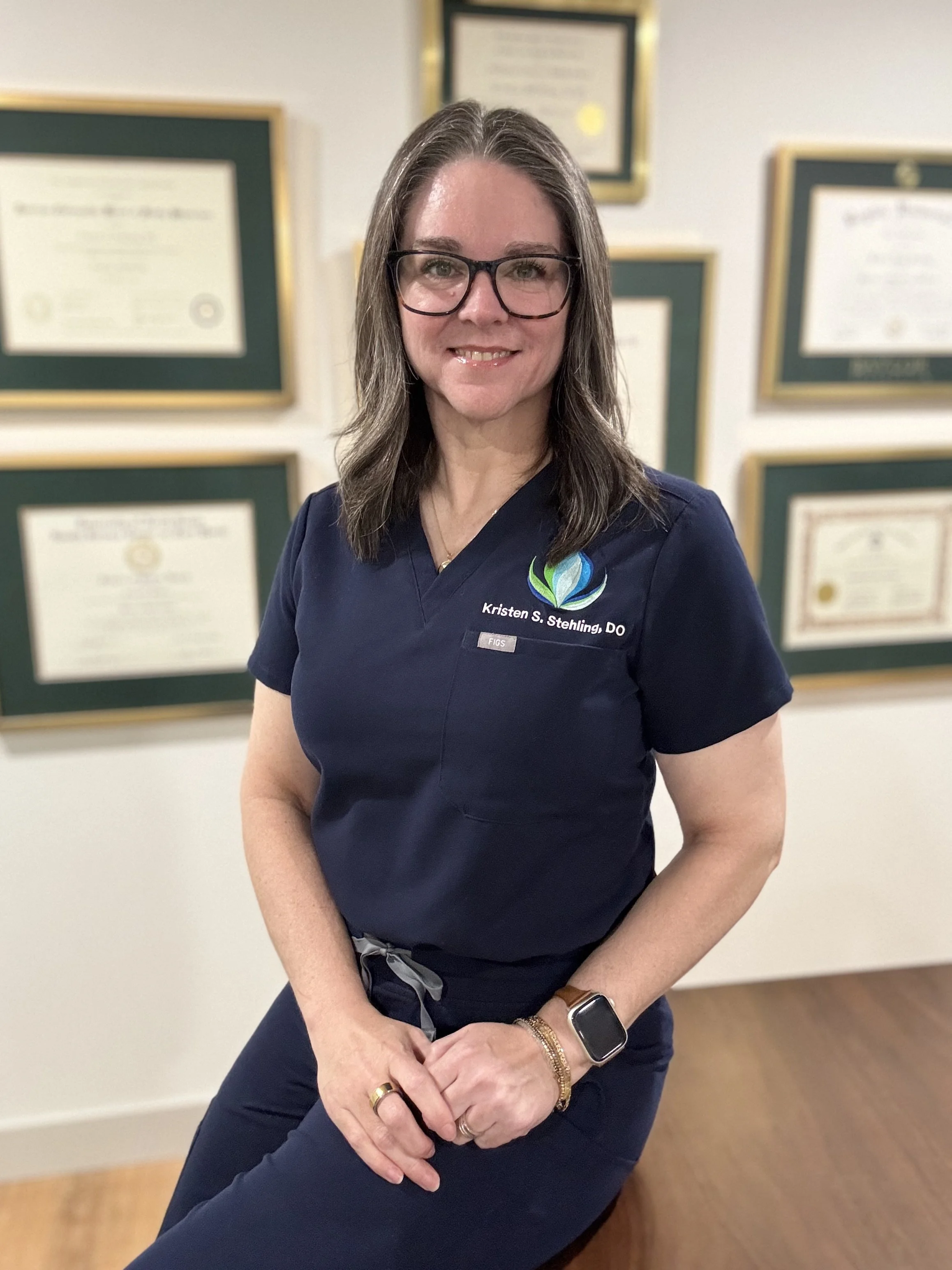 A woman with long brown hair and glasses wearing navy scrubs standing in front of framed certificates or diplomas on a wall.