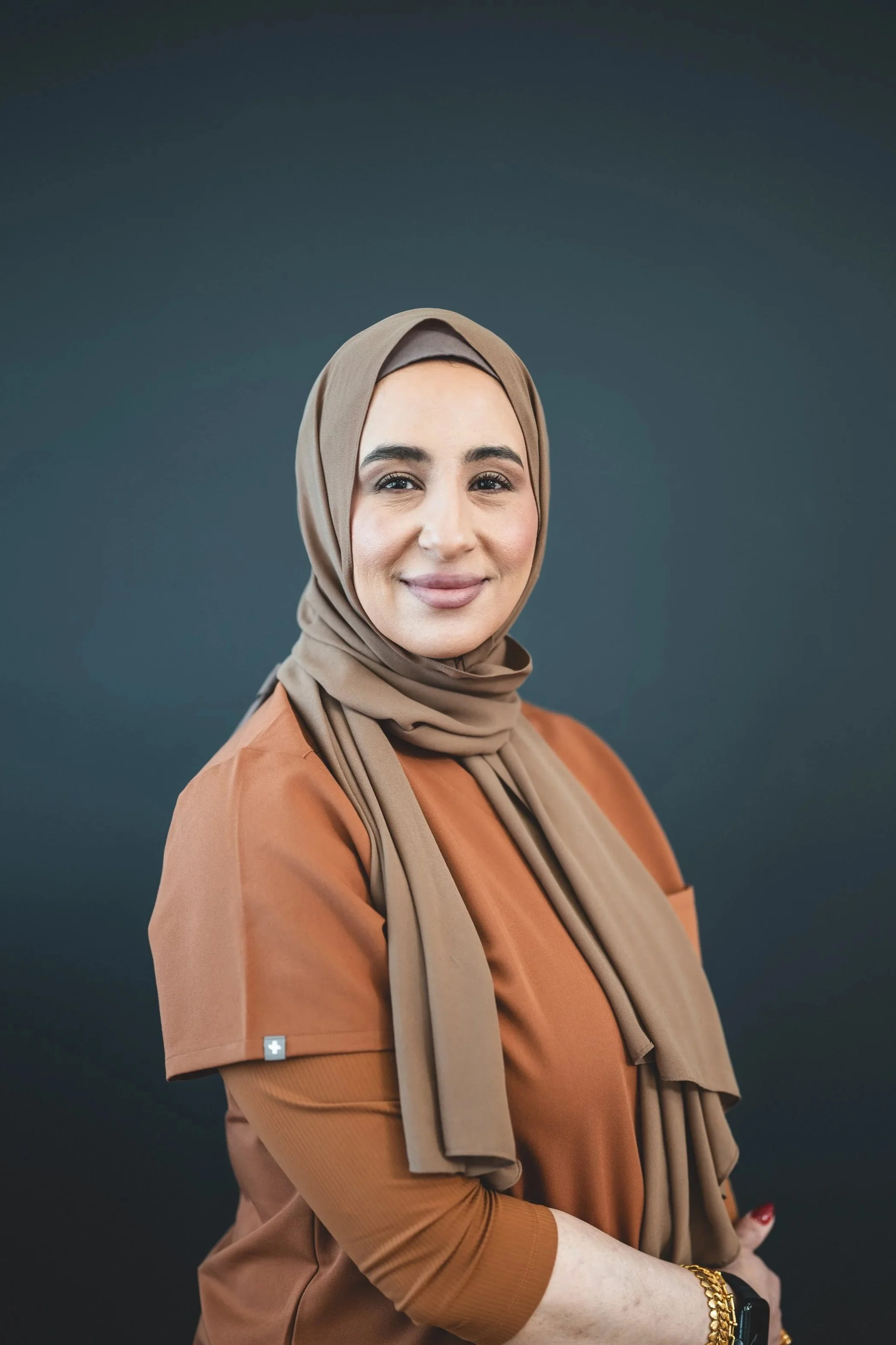 A woman wearing a beige hijab and a matching brown jacket, smiling softly against a dark background.