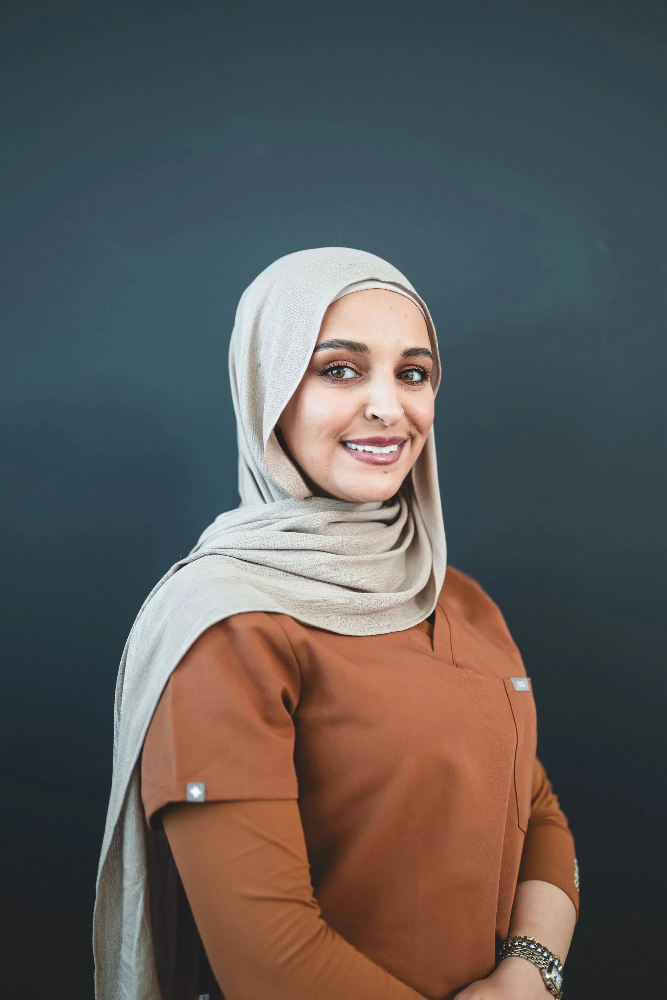 A woman wearing a beige hijab and a brown top, smiling and posing against a dark background.
