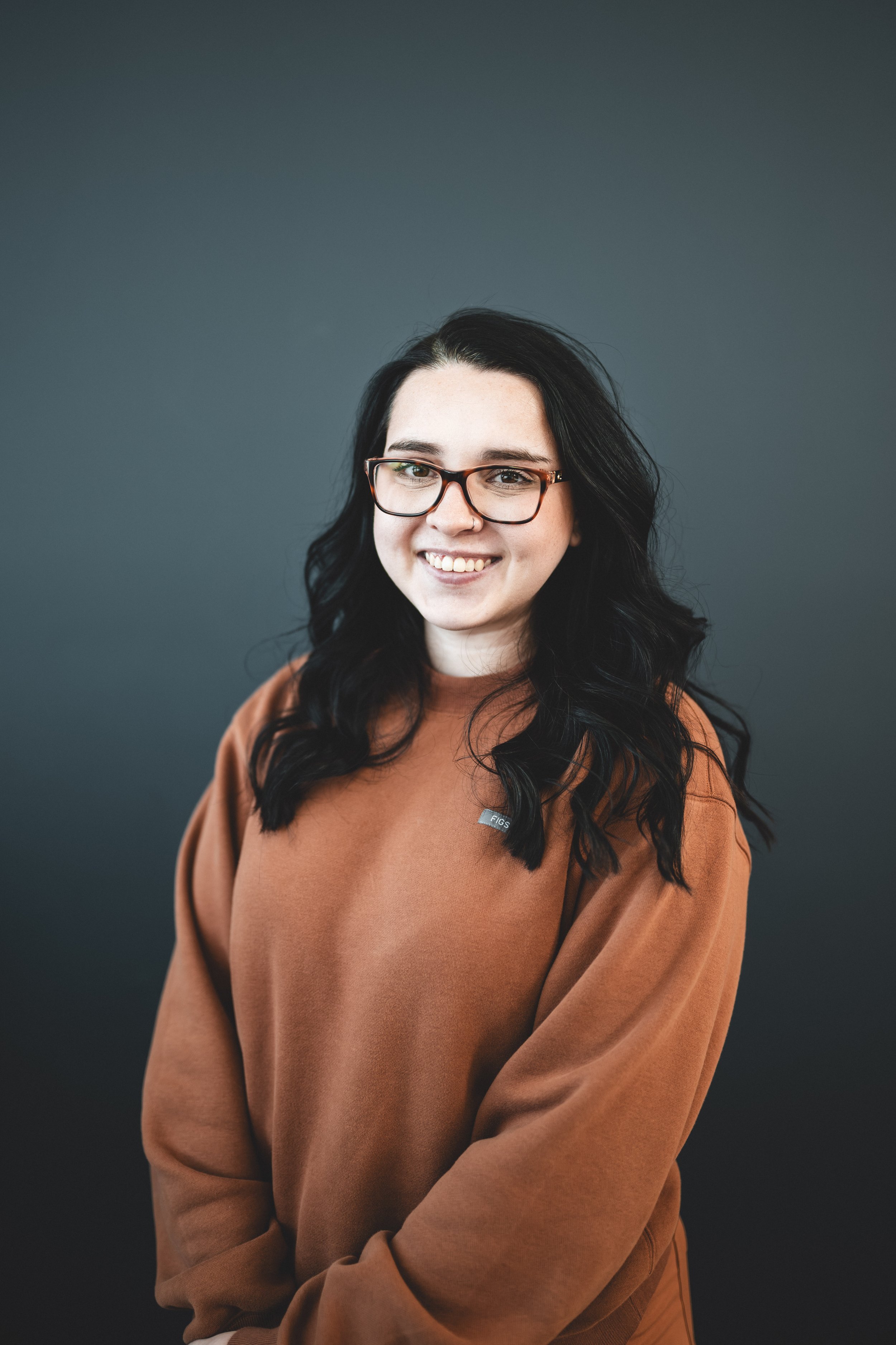 A woman with long black hair, glasses, and a warm smile, wearing a brown sweater, stands in front of a plain dark background.