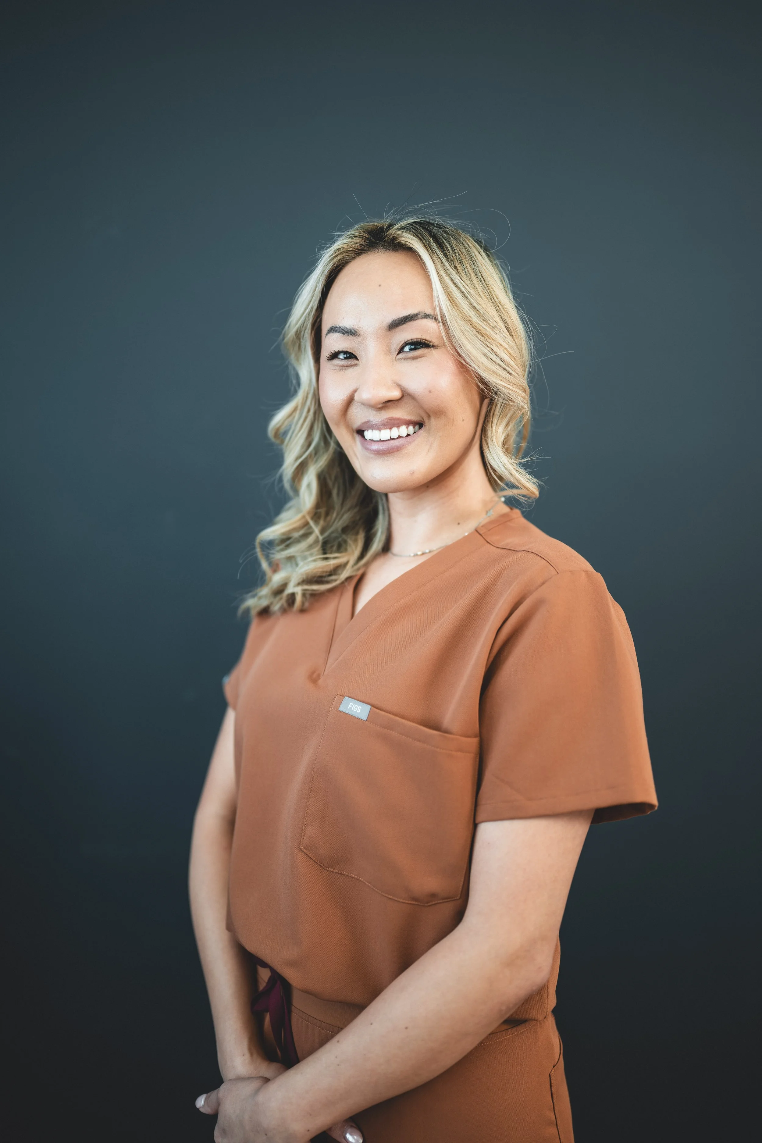 Portrait of a smiling woman with wavy blonde hair, wearing a brown scrub top, standing against a dark background.
