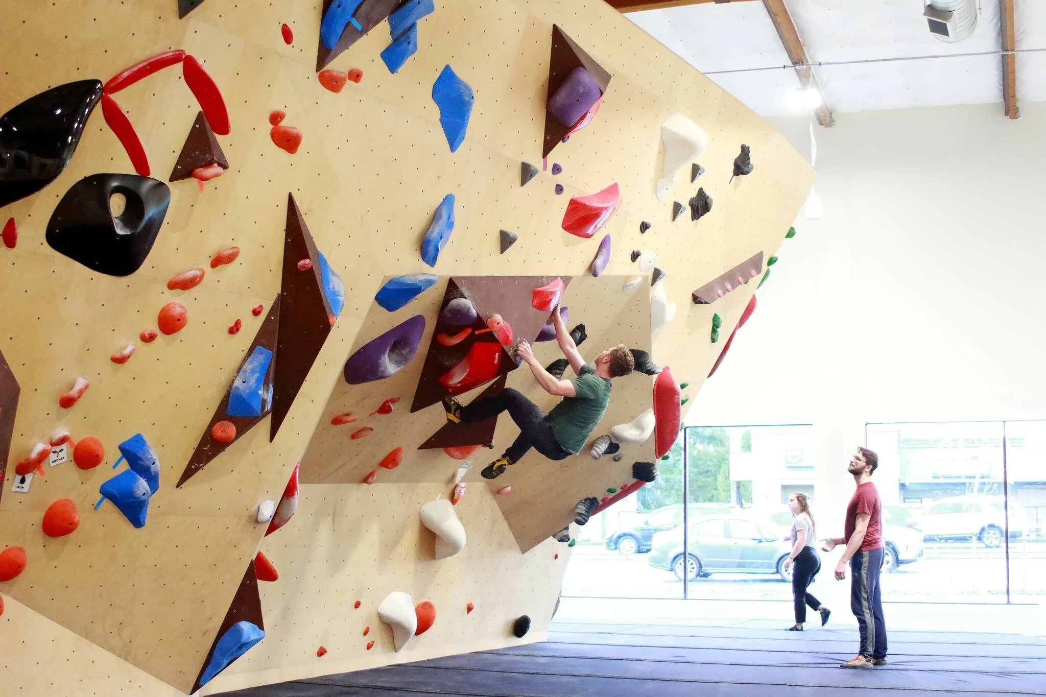 Indoor bouldering gym with various colorful climbing holds on a large wooden climbing wall. Several people are engaged in climbing or observing, and large windows let in natural light.
