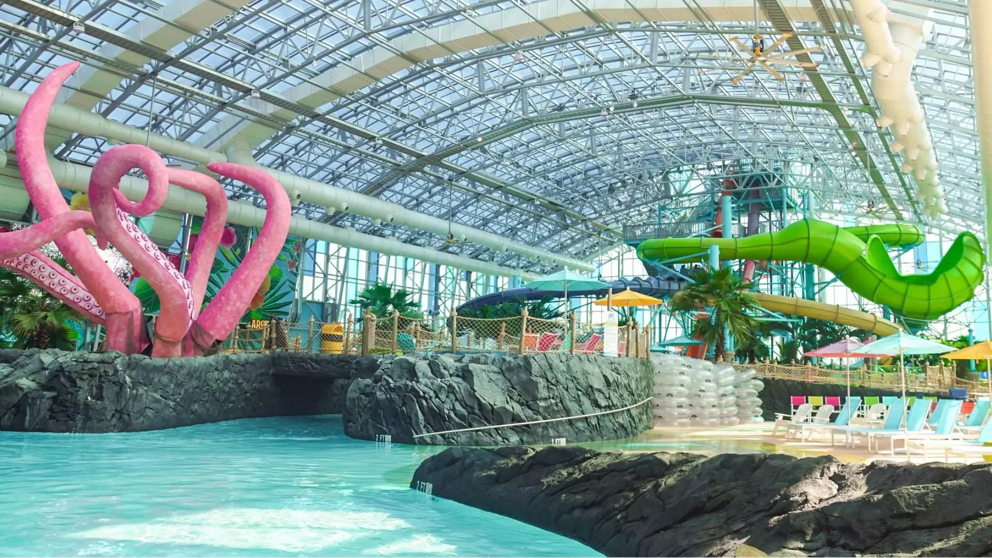 Indoor water park with large water slides, a lazy river, and pool area under a glass roof, decorated with tropical plants and colorful umbrellas.