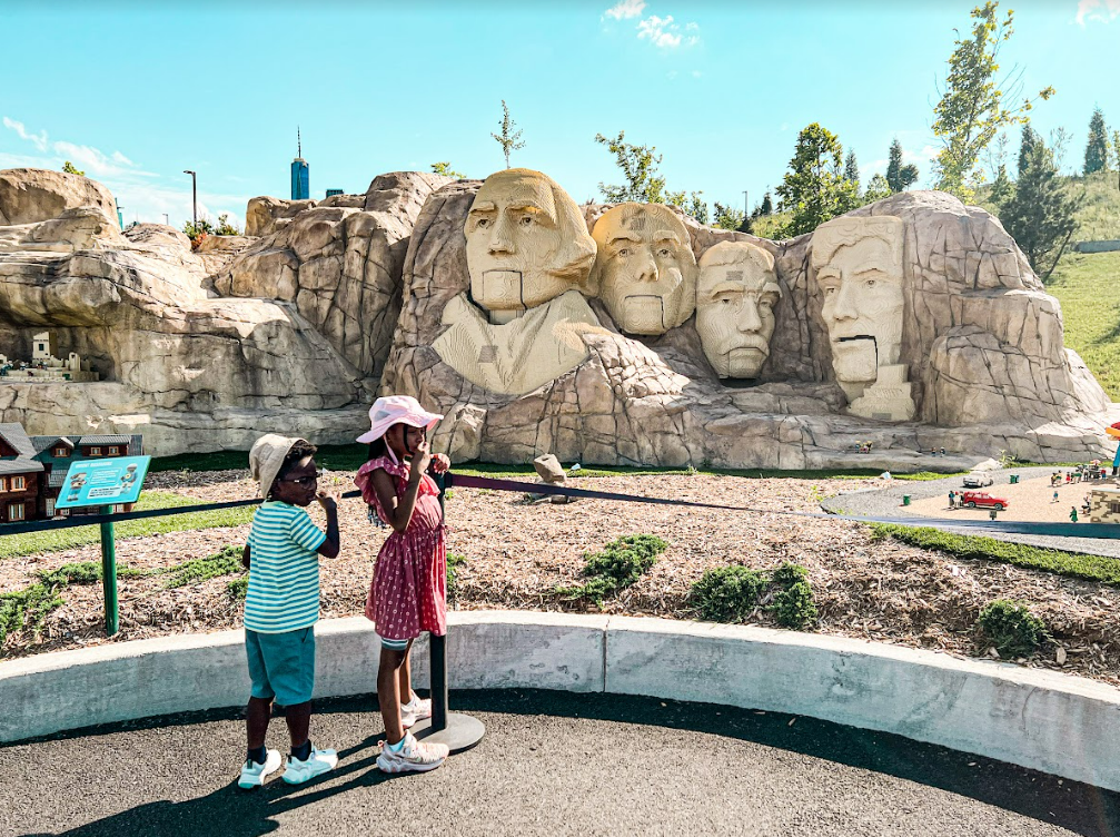 Two children, one girl and one boy, standing in front of a miniature replica of Mount Rushmore at a park, with a blue sky and trees in the background.
