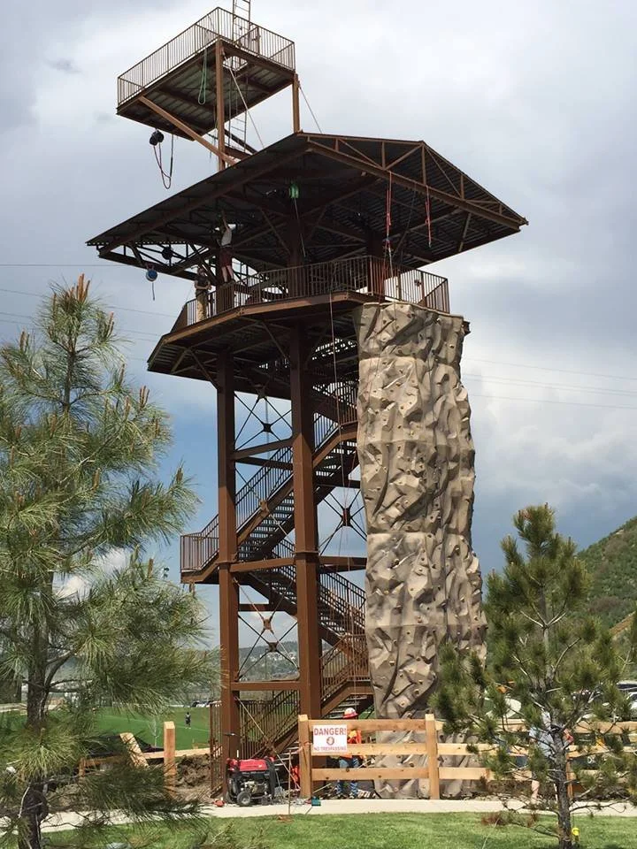 An outdoor observation tower with a rock climbing wall attached, set against a cloudy sky, surrounded by trees and a grassy area.