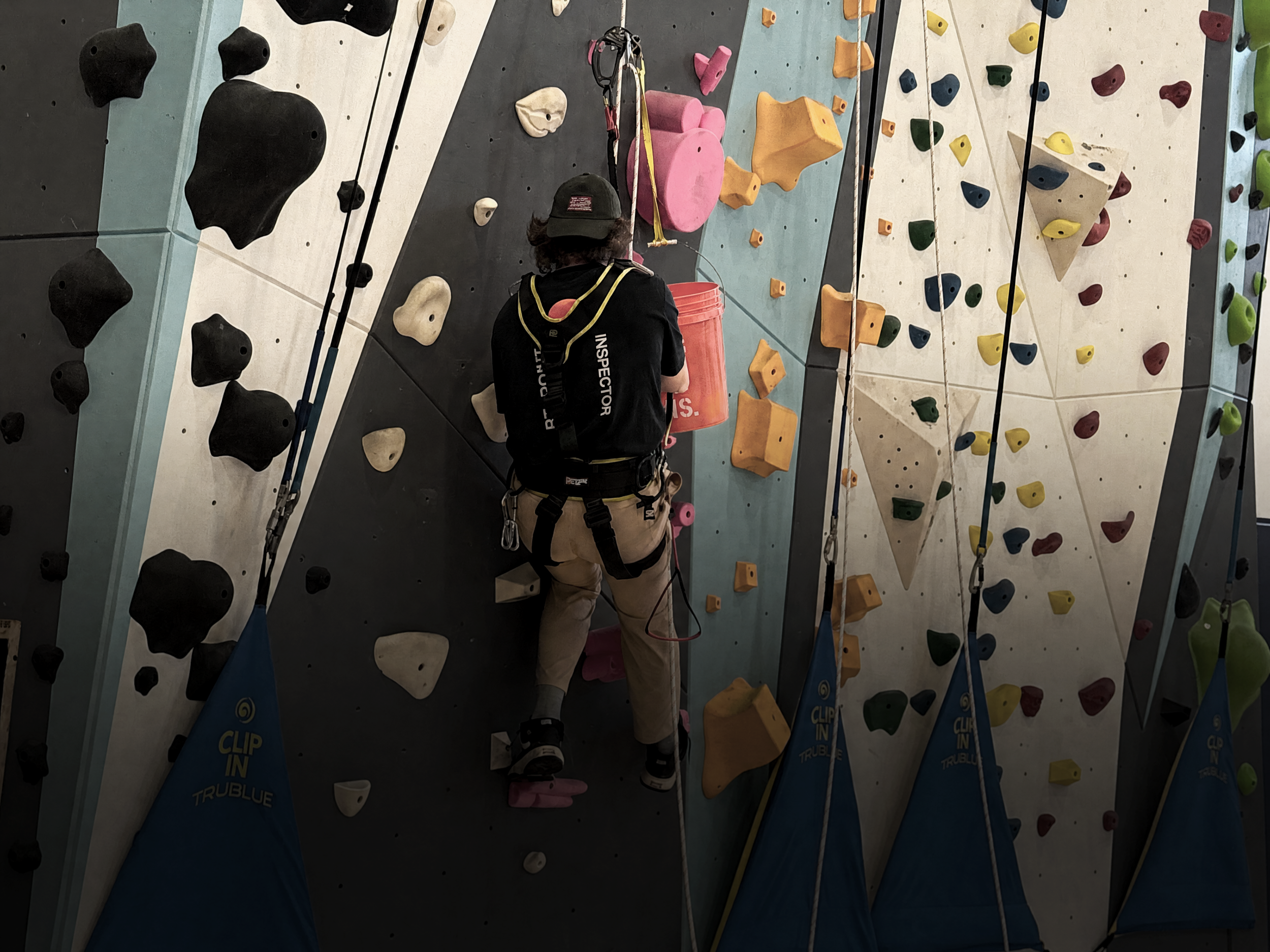 A person wearing a black helmet and harness climbing an indoor boulder wall with colorful holds in black, white, pink, orange, yellow, green, and blue.