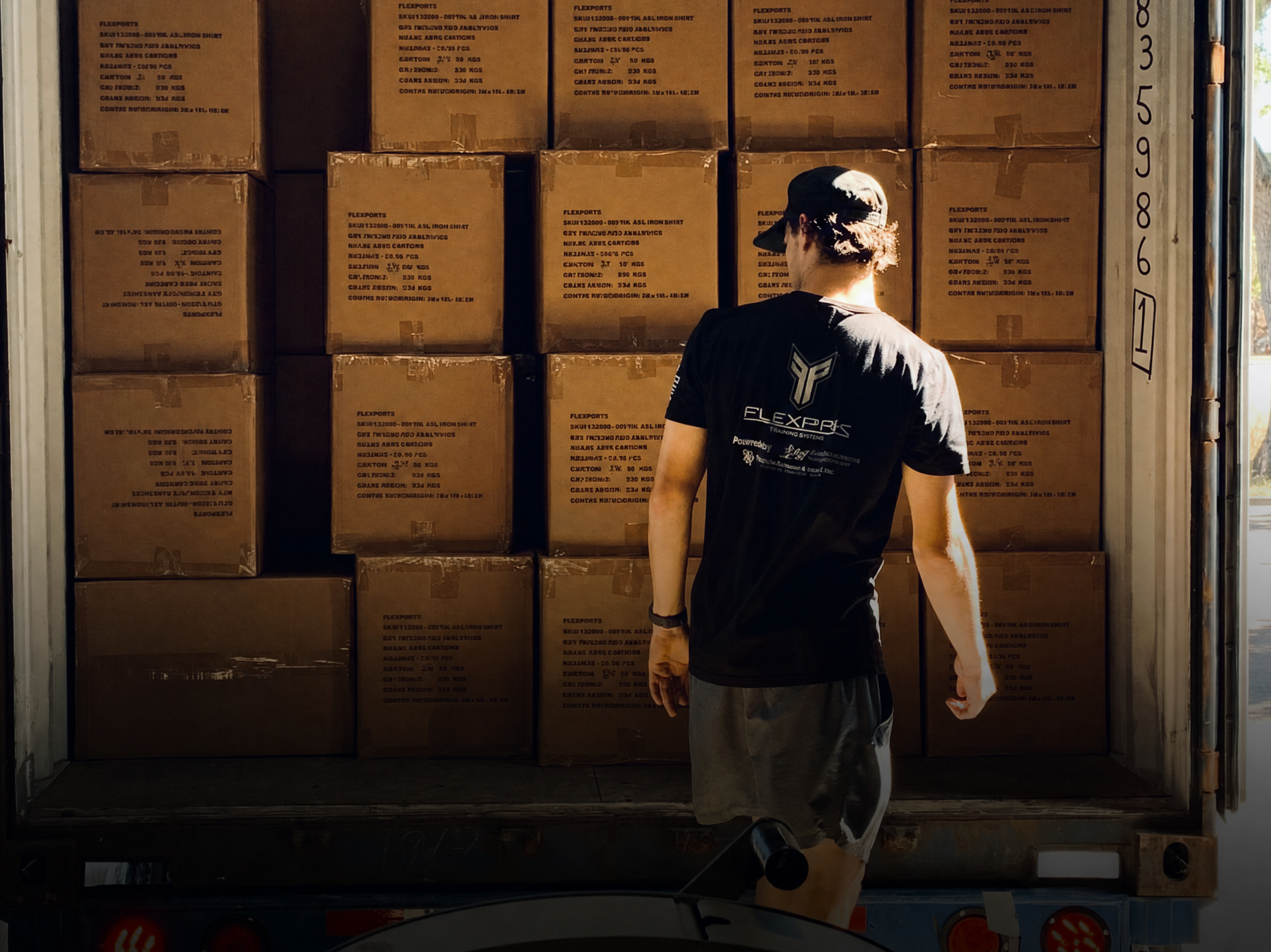 A person with curly hair wearing a black cap and a black T-shirt with a logo and text, standing in front of stacked cardboard boxes inside a truck or storage area.