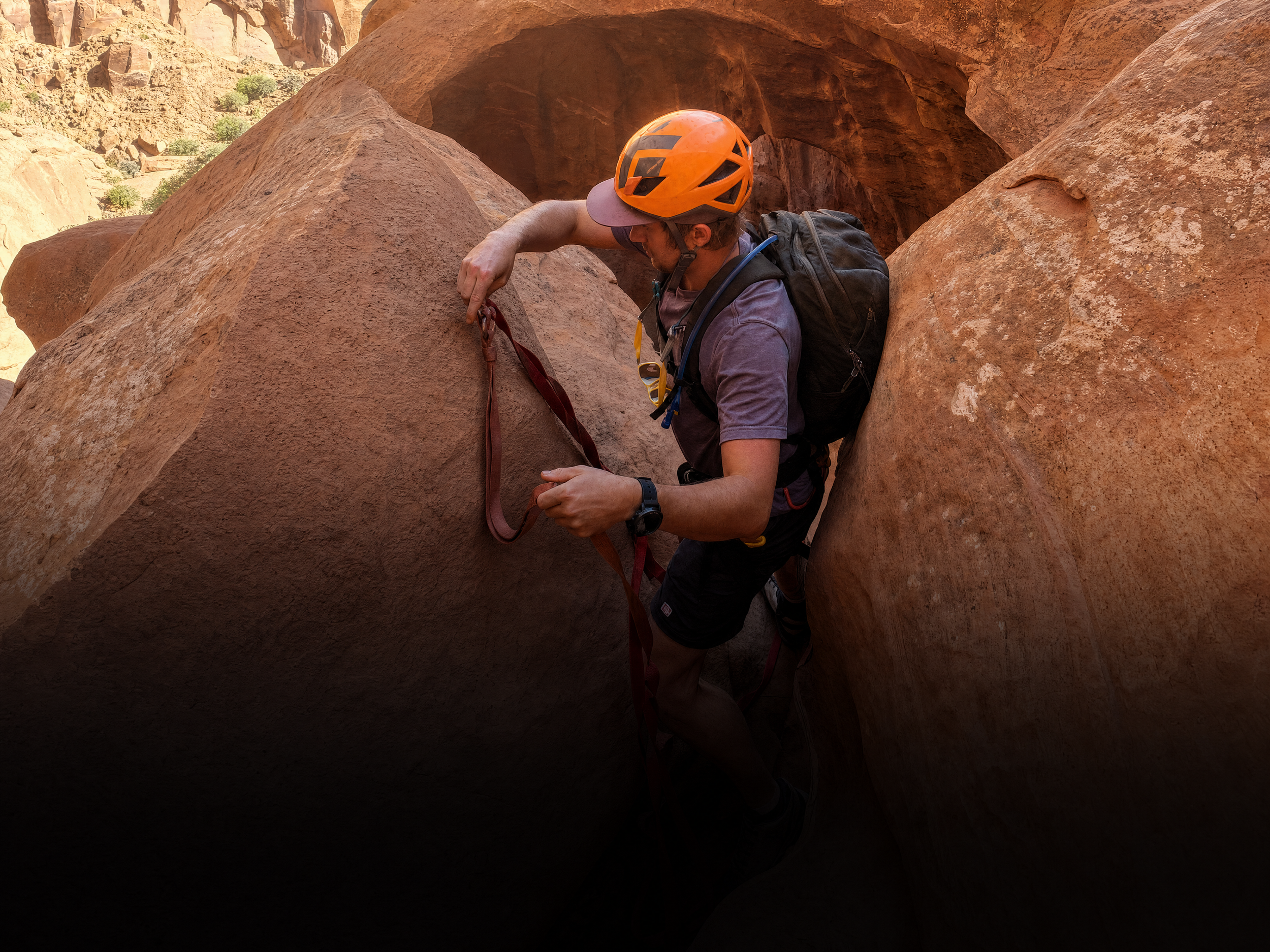 A man wearing a helmet and backpack climbing through a narrow gap between large red rocks in a desert landscape.
