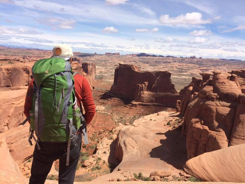 A hiker with a large green backpack and a beige cap walking through a canyon landscape of red rock formations and a partly cloudy sky.