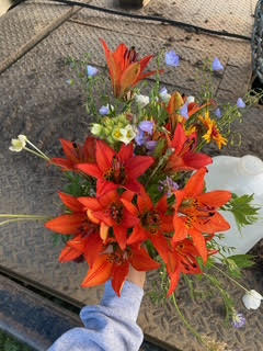 A bouquet of vibrant orange lilies with smaller purple, white, and yellow flowers, held by a person wearing a gray glove, outdoors on a glass table.