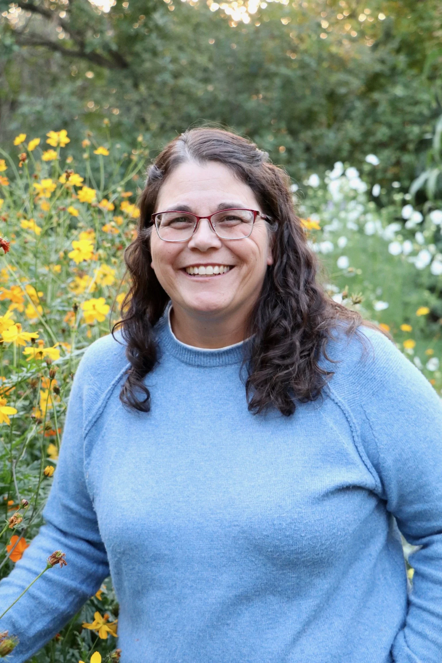 A woman with glasses and long curly dark hair smiling outdoors, standing among yellow flowers, with green trees in the background.