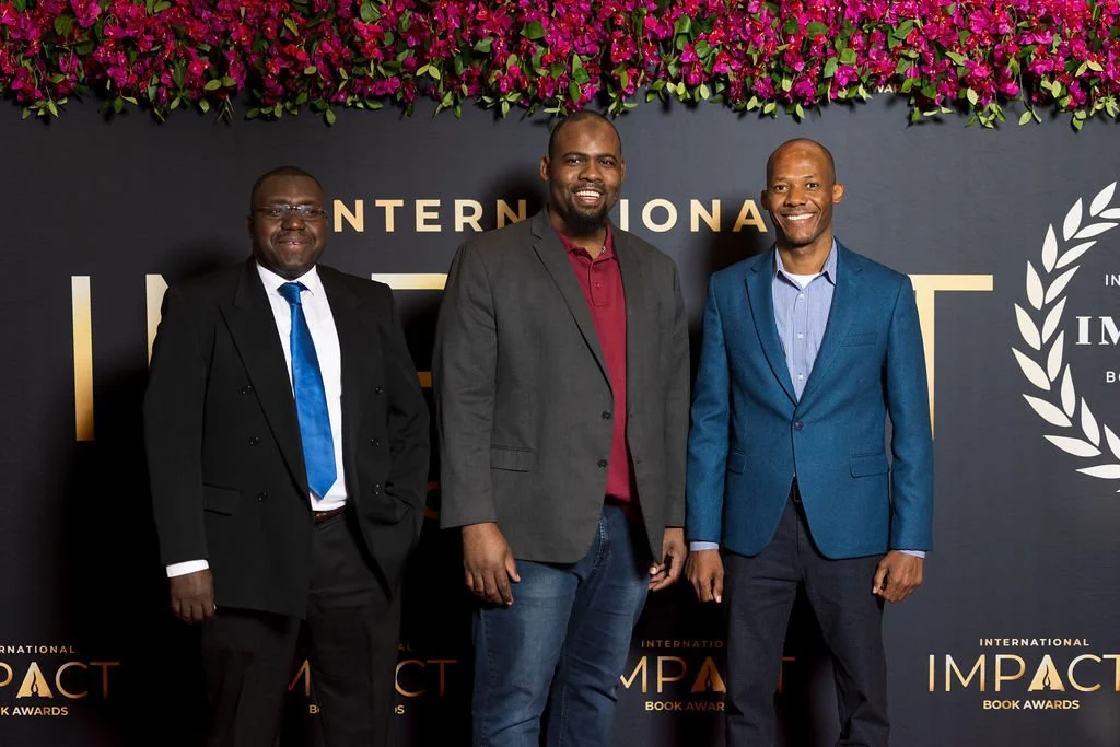 Three men in suits standing in front of a black background with pink flowers on top, at an international impact book awards event.