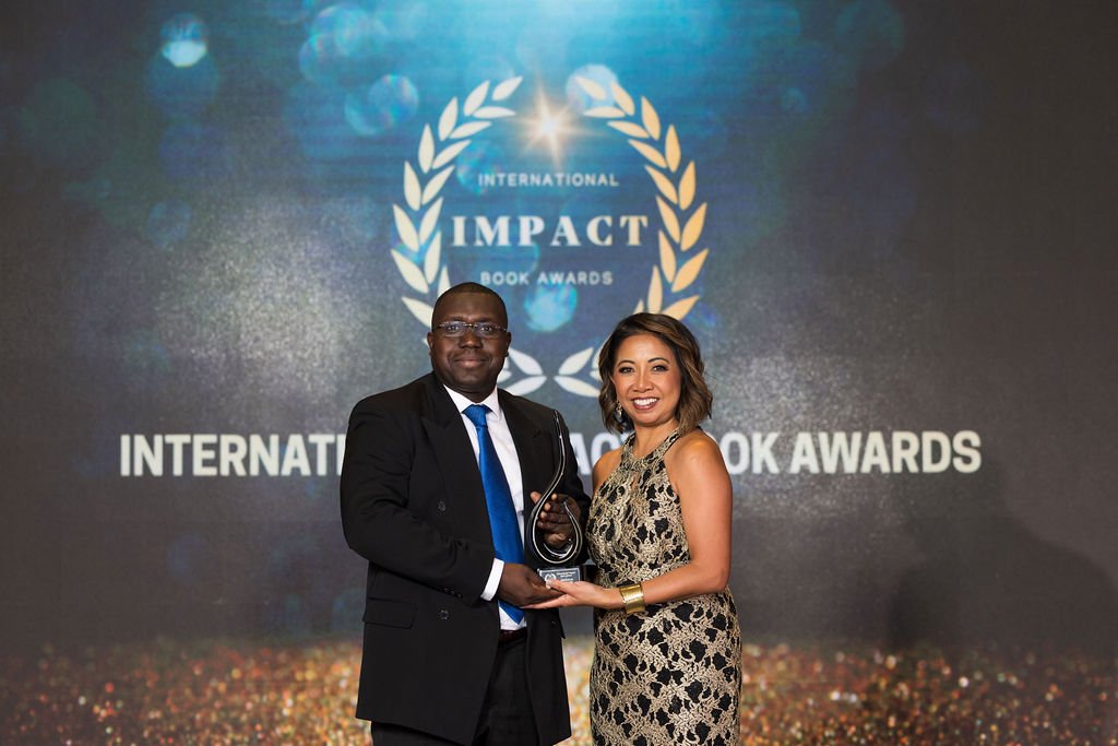 A man in a black suit and blue tie and a woman in a gold and black dress holding an award on stage at the International Impact Book Awards, with a large screen displaying the event's logo behind them.