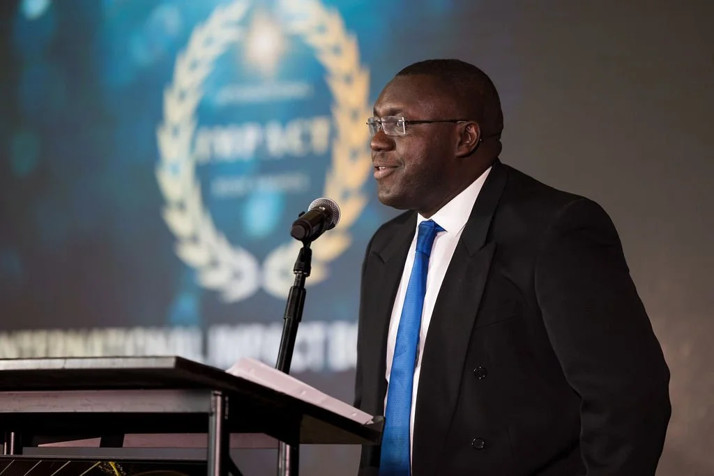 A man in a black suit, white shirt, and blue tie speaking at a podium with a microphone, behind a blurred logo on a screen.