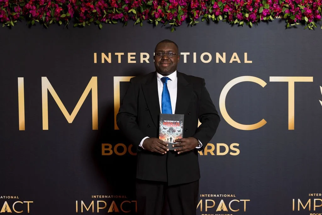 A man in a black suit and blue tie standing on stage at the International IMPACT Book Awards, holding a book titled 'The Realm of the Evil.' Behind him is a black backdrop with gold and white text that reads 'INTERNATIONAL IMPACT BOOK AWARDS,' decorated with purple flowers at the top.