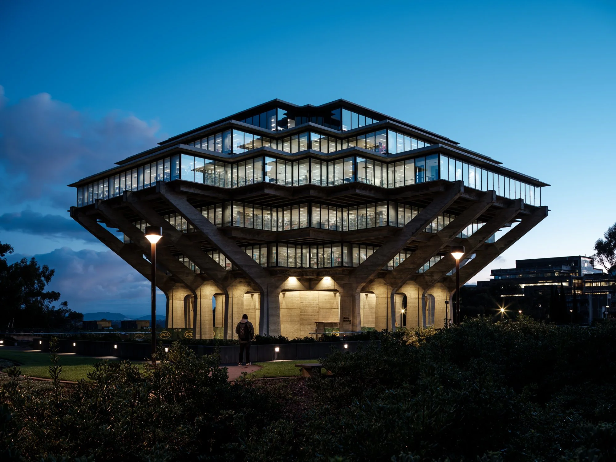 A modern, glass-walled building with multiple stories illuminated from within, supported by large concrete pillars with a unique architectural design, set against a twilight sky. A person stands in the foreground.