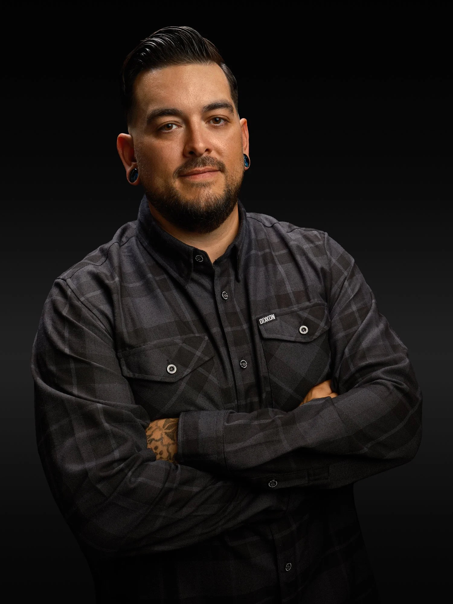 Portrait of a man with dark hair slicked back, facial hair, and stretched earlobes wearing a black and gray checkered shirt with arms crossed against a black background.