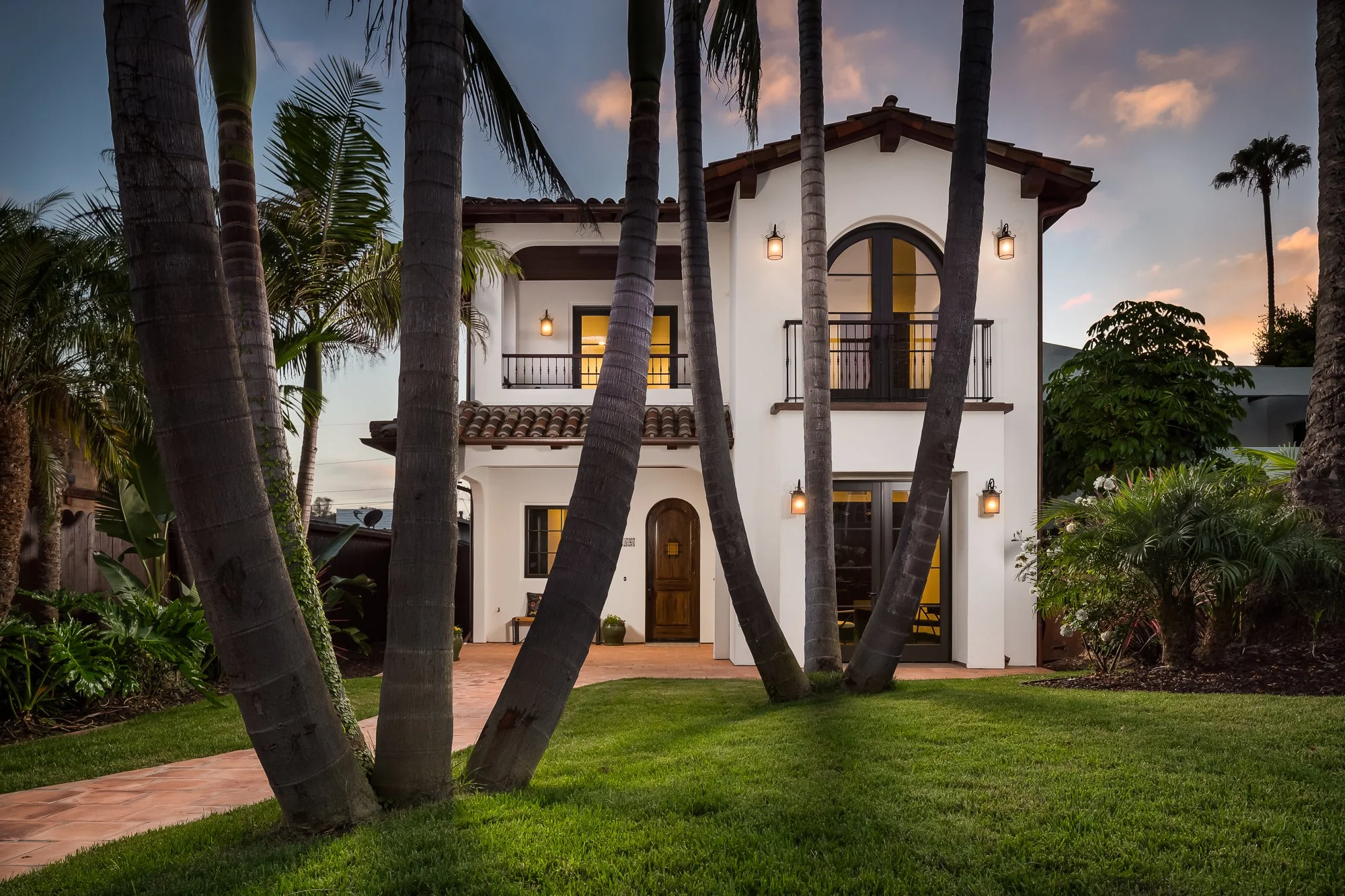 A modern two-story house with white exterior walls, large arched window, and balcony, surrounded by tall palm trees and lush greenery during sunset.