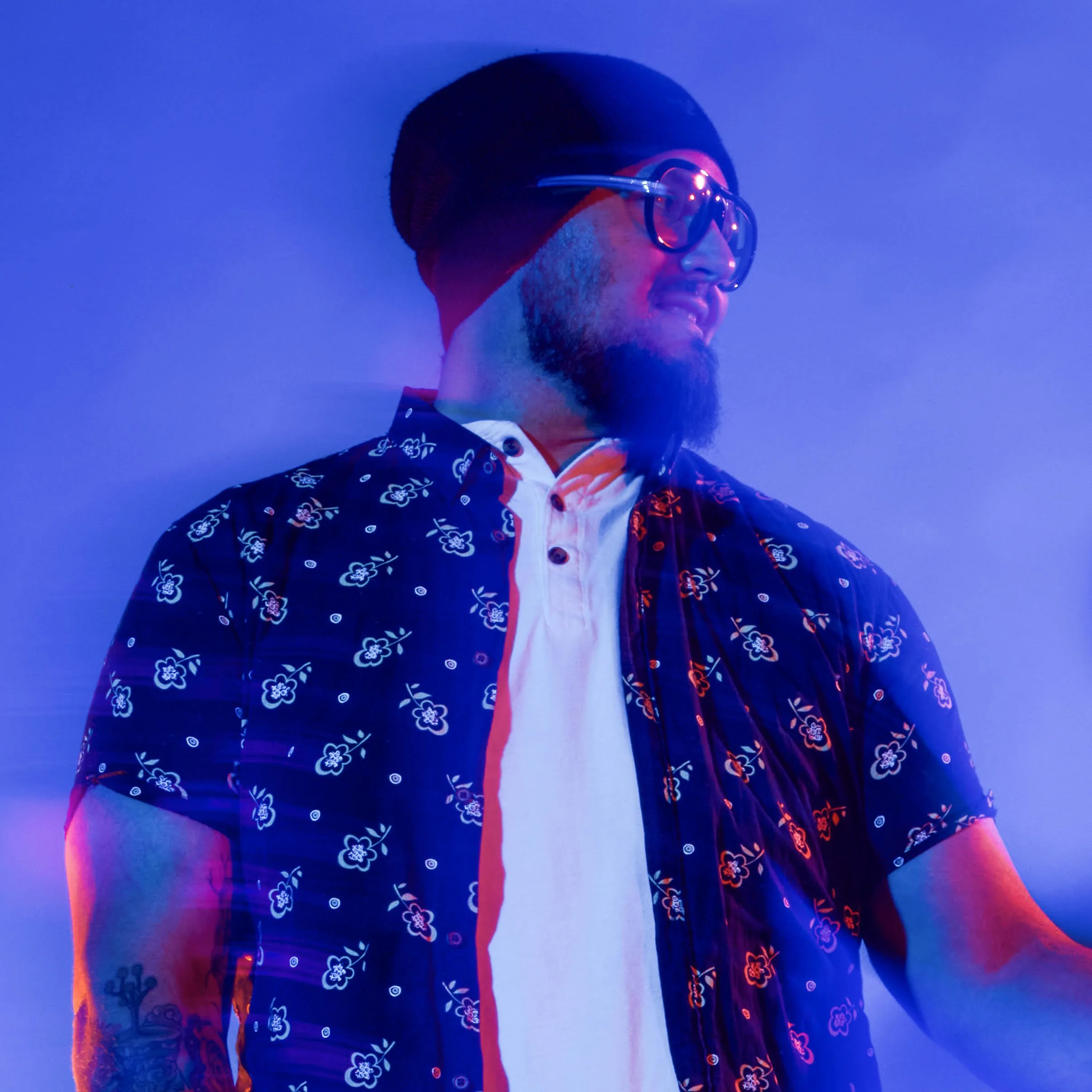 A man with a beard and sunglasses is smiling against a blue lit background, wearing a black shirt with colorful floral patterns.