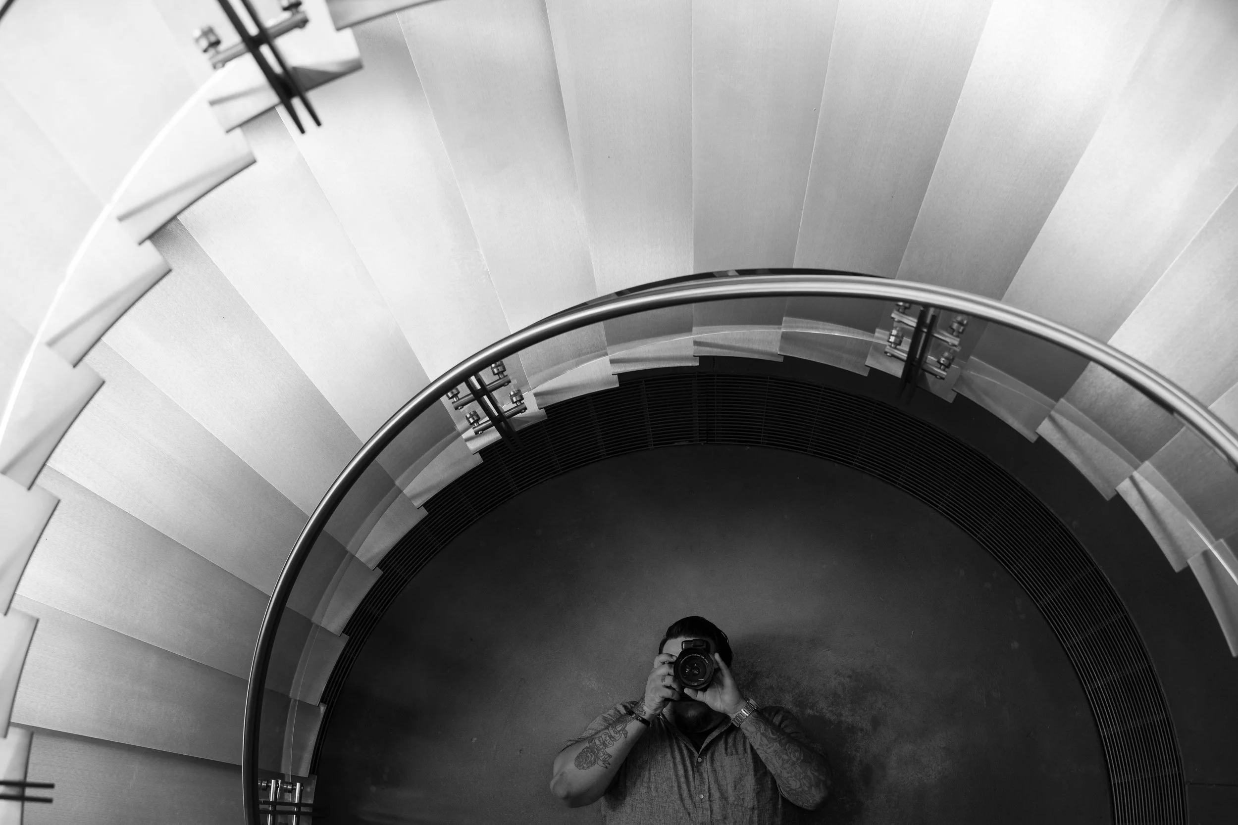 Black and white photo of a person taking a picture of themselves using a smartphone in a large, spiral staircase with a circular railing, viewed from above.