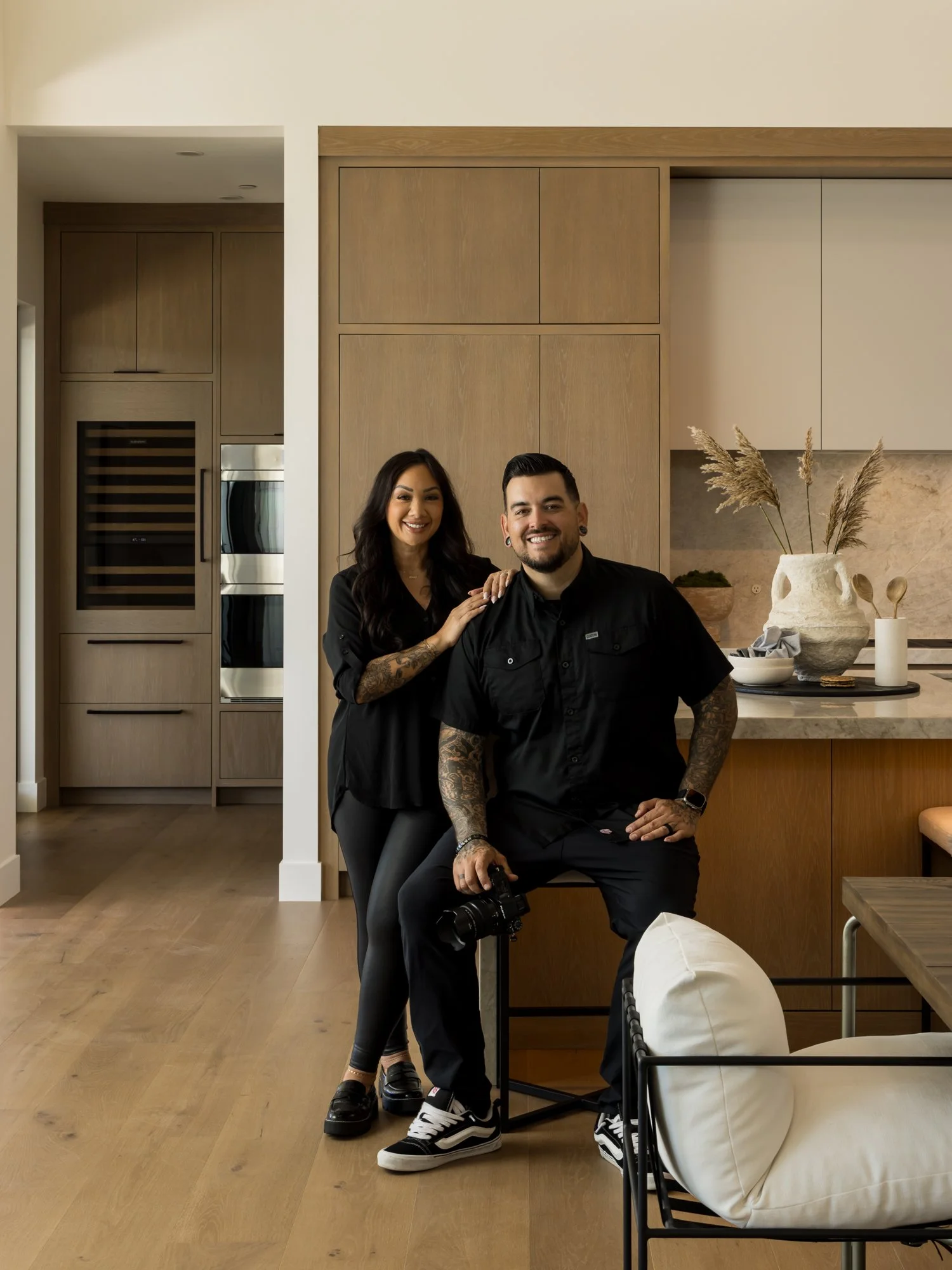 A smiling woman with dark wavy hair and a tattoo on her arm standing behind a seated man with short dark hair, a beard, and tattoos on his arms, both dressed in black, inside a modern kitchen with wooden cabinetry, a marble countertop, decorative vases, and kitchen utensils.