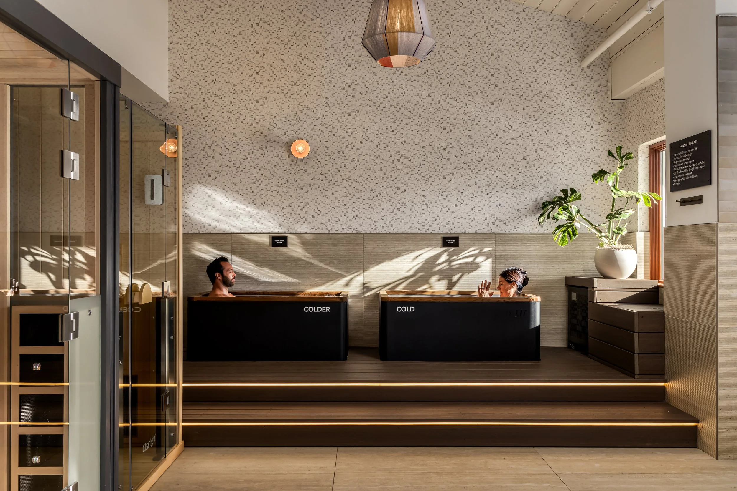 People enjoying a bath in a modern spa setting with wooden tubs labeled 'COLDER' and 'COLD,' a potted plant, a textured wall, and ambient lighting.