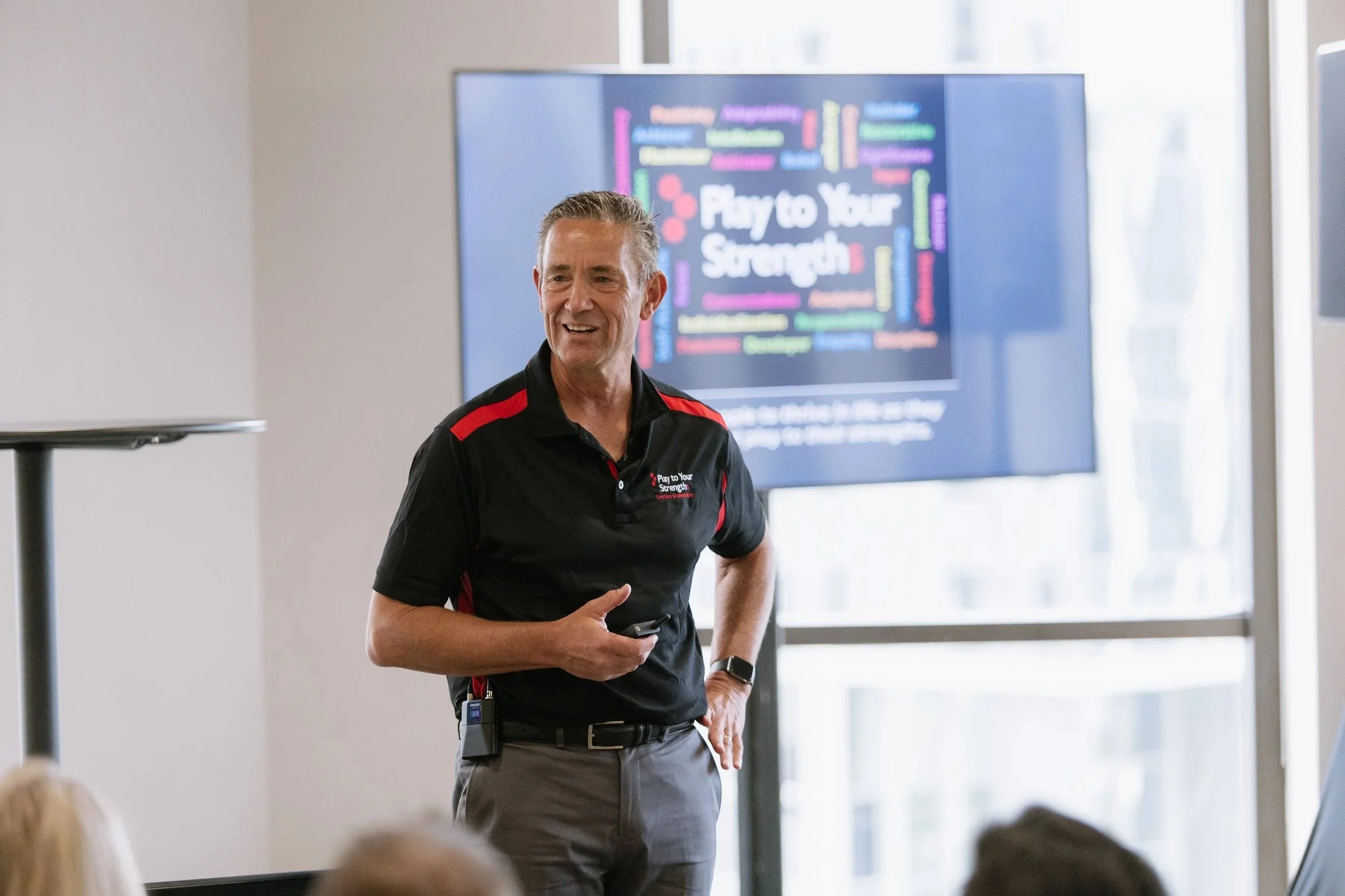 A man in a black polo shirt with red accents speaking or presenting at an event. Behind him, a large screen displays a colorful slide with the phrase 'Play to Your Strength,' and the audience is visible in the foreground.