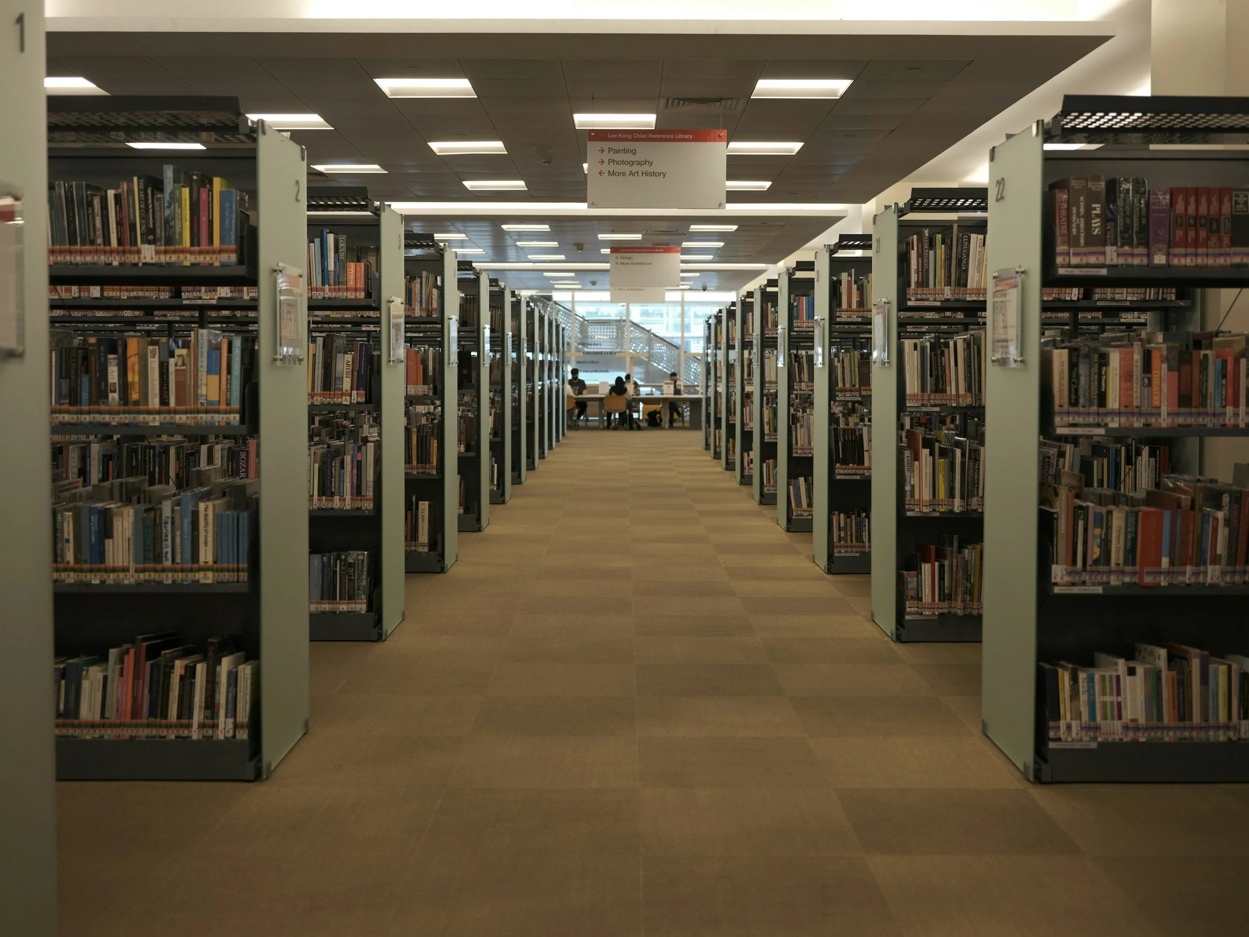 Rows of bookshelves filled with books in a library, with signs hanging from the ceiling for painting, photography, and art history sections.