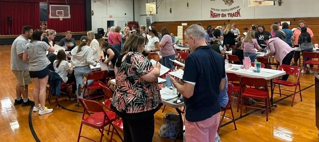 People gather around tables in a gymnasium, participating in an event with food and drinks, some standing and others seated, with a basketball hoop and gym equipment visible in the background.