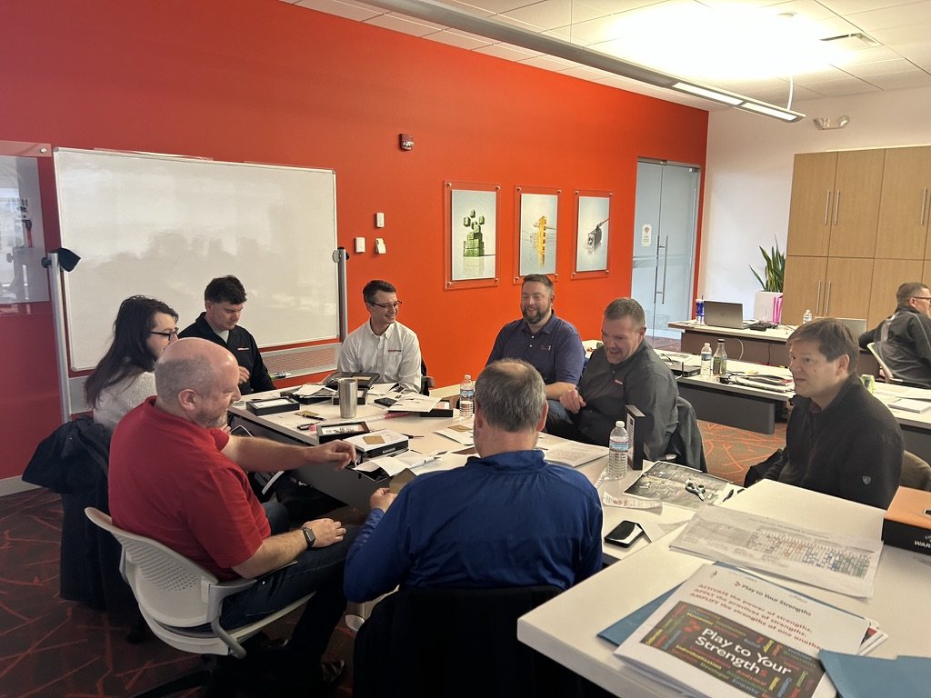 Group of eight people sitting around a conference table in a brightly lit meeting room with orange walls, whiteboard, and framed artwork, engaged in discussion with water bottles and documents on the table.