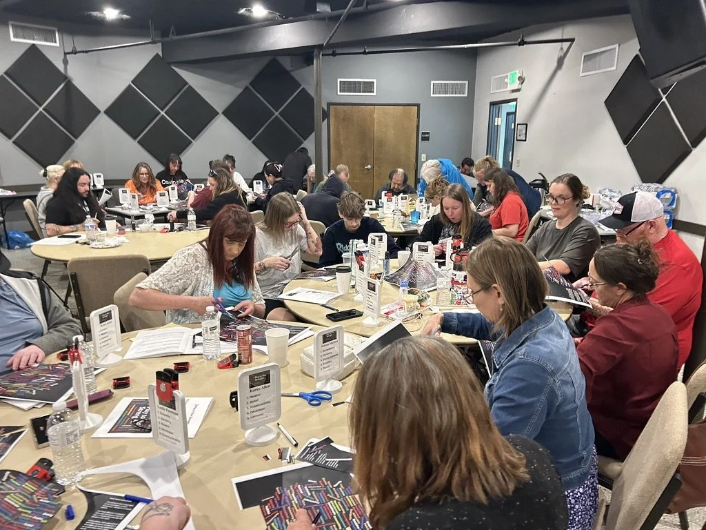 People sitting around tables in a room, engaging in an activity involving small objects, papers, and tools, with some using scissors and counters, in a setting with soundproofing panels on the walls.