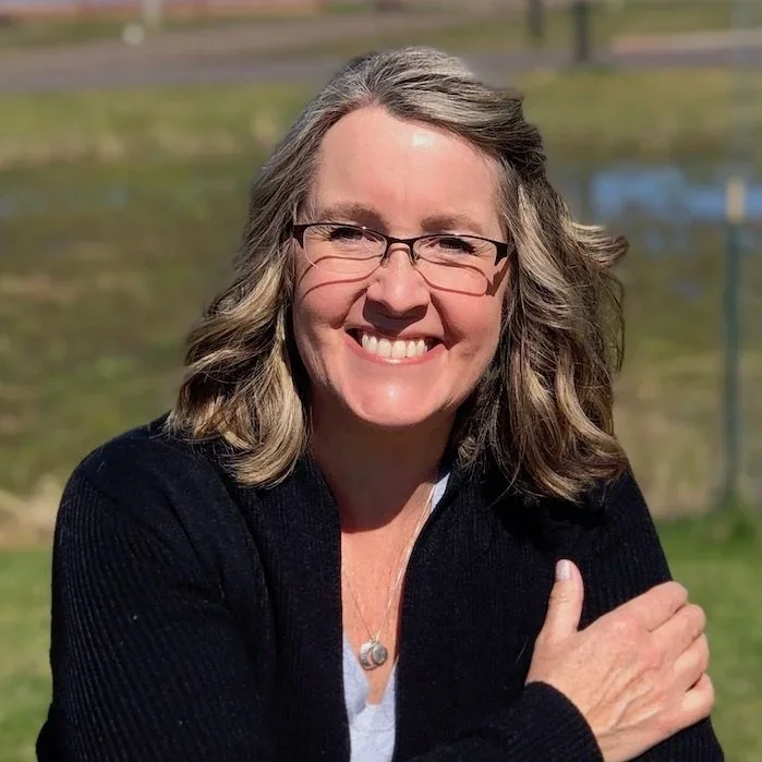 A woman with shoulder-length wavy hair, glasses, and a big smile, standing outdoors with a blurred background of grass and trees.