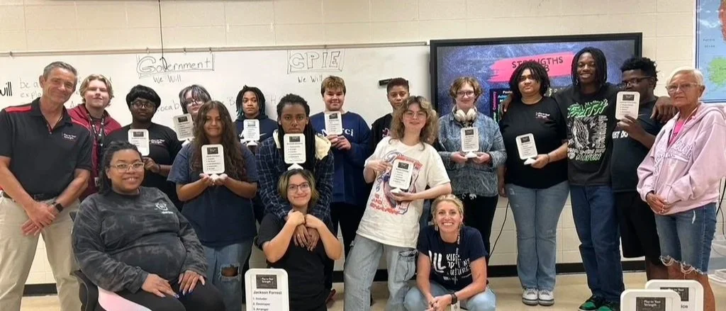 Group of students and teachers posing in a classroom, holding awards or books, in front of a whiteboard and a digital display.