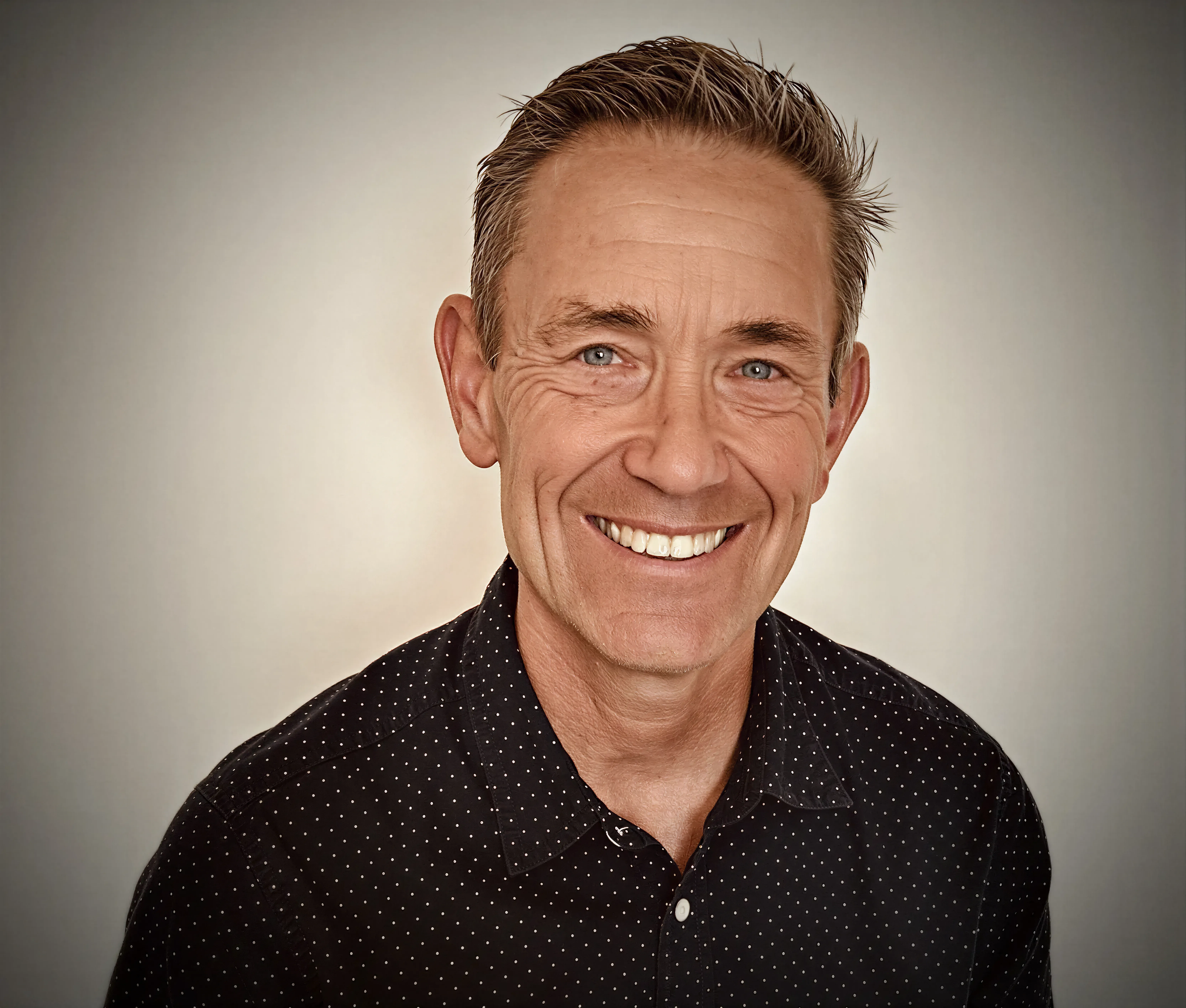 Close-up of smiling middle-aged man with brushed-back hair, wearing a black shirt with white polka dots, against a plain light background.