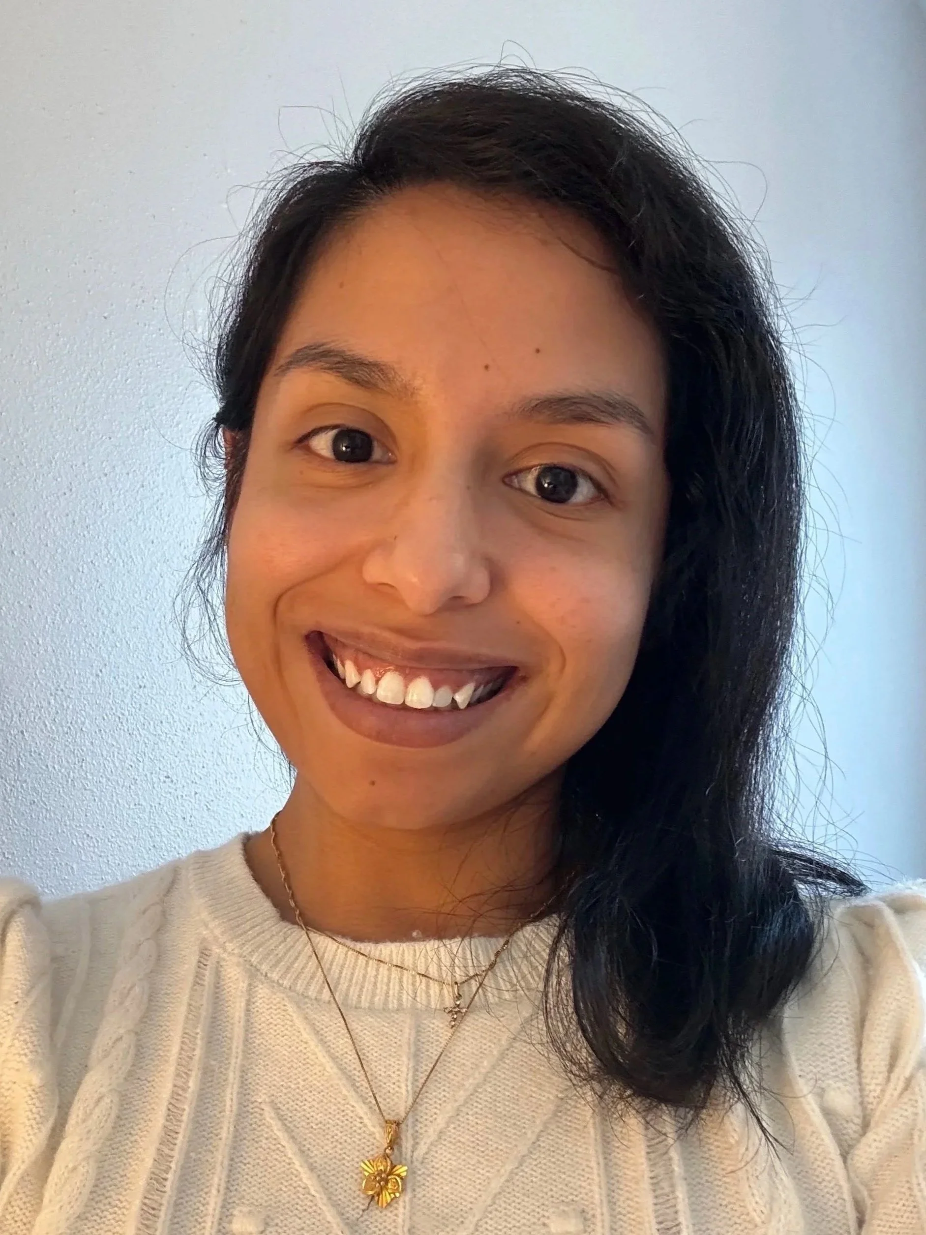 A close-up photo of a woman smiling, wearing a cream-colored sweater and a gold necklace with a floral pendant, with dark hair and clear skin, standing against a plain light blue background.