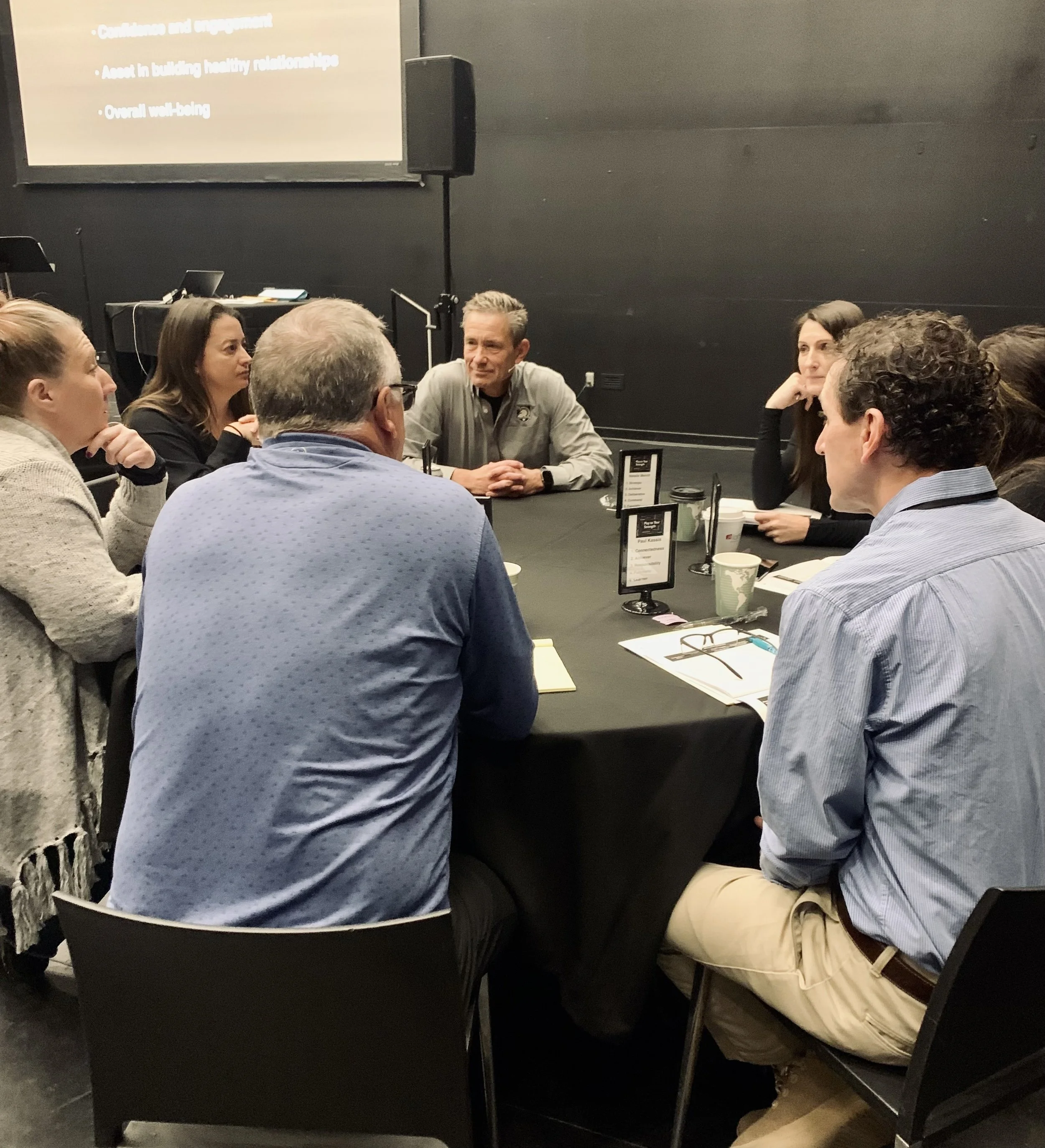 A group of people sitting at a table in a dark room, engaged in a discussion or meeting. There are name tags, notebooks, and coffee cups on the table, with a presentation slide visible on the wall behind them.