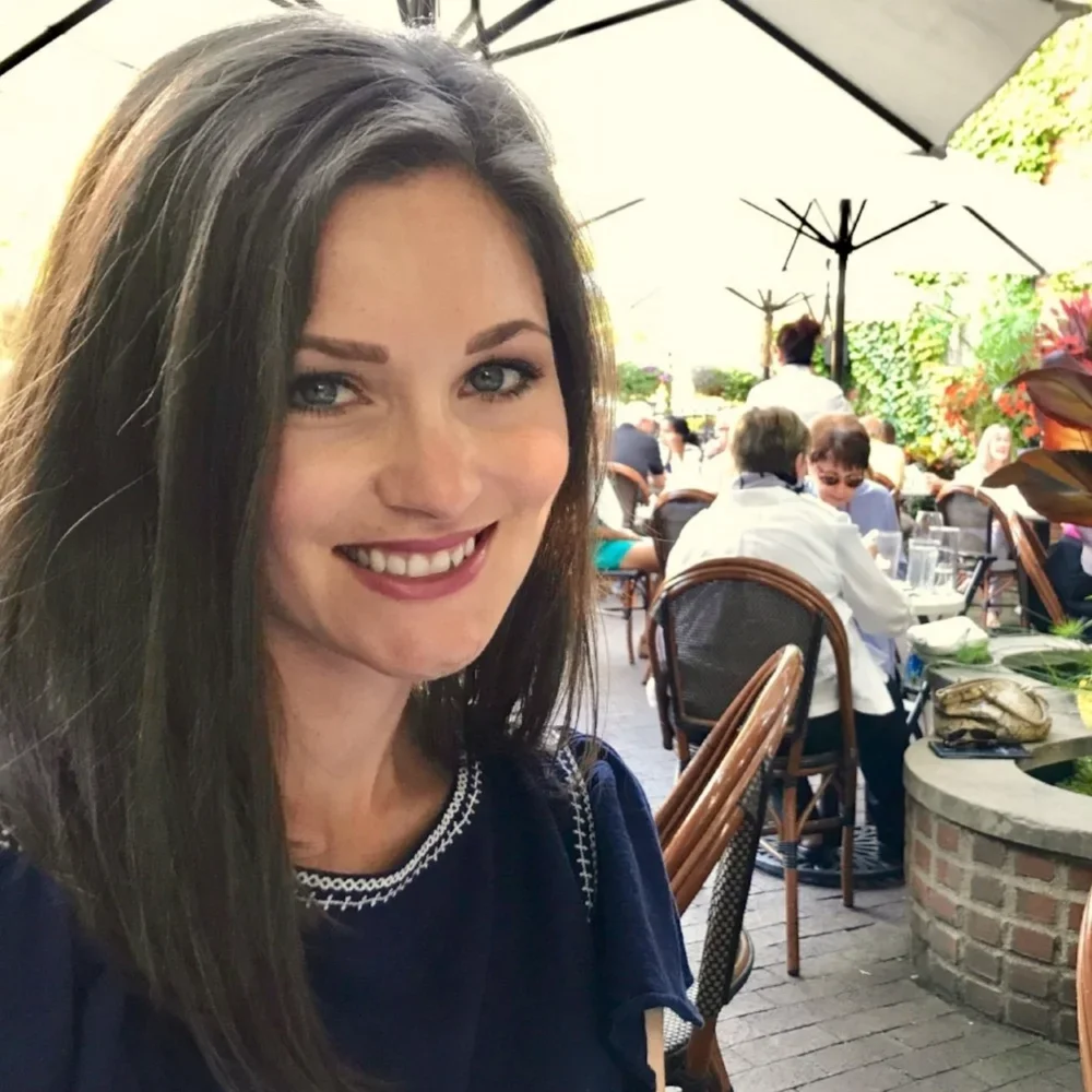 A woman with long dark hair and a dark top smiling at a restaurant or cafe with outdoor seating, tables, and umbrellas, filled with people dining.