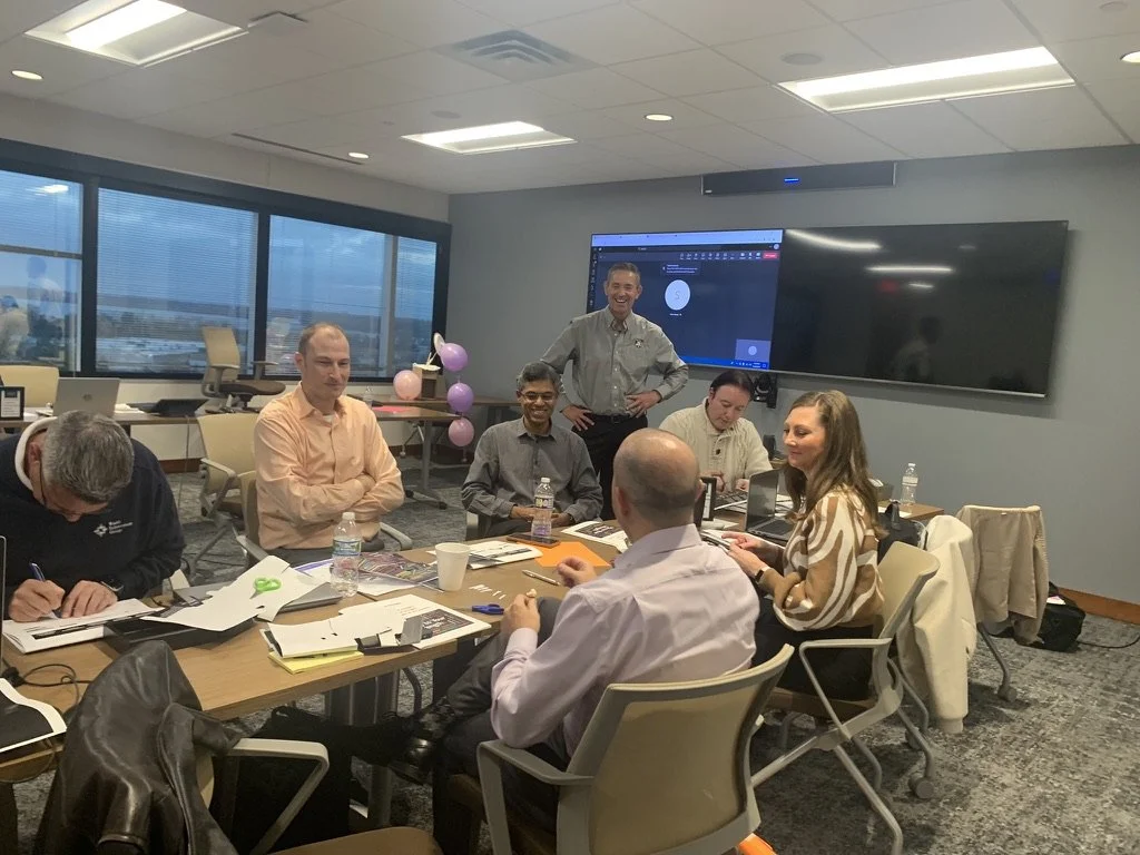 A group of eight people in a conference room, some sitting at a table and one standing, with a large window showing an outdoor view, balloons, laptops, and papers on the table, and a TV screen on the wall.