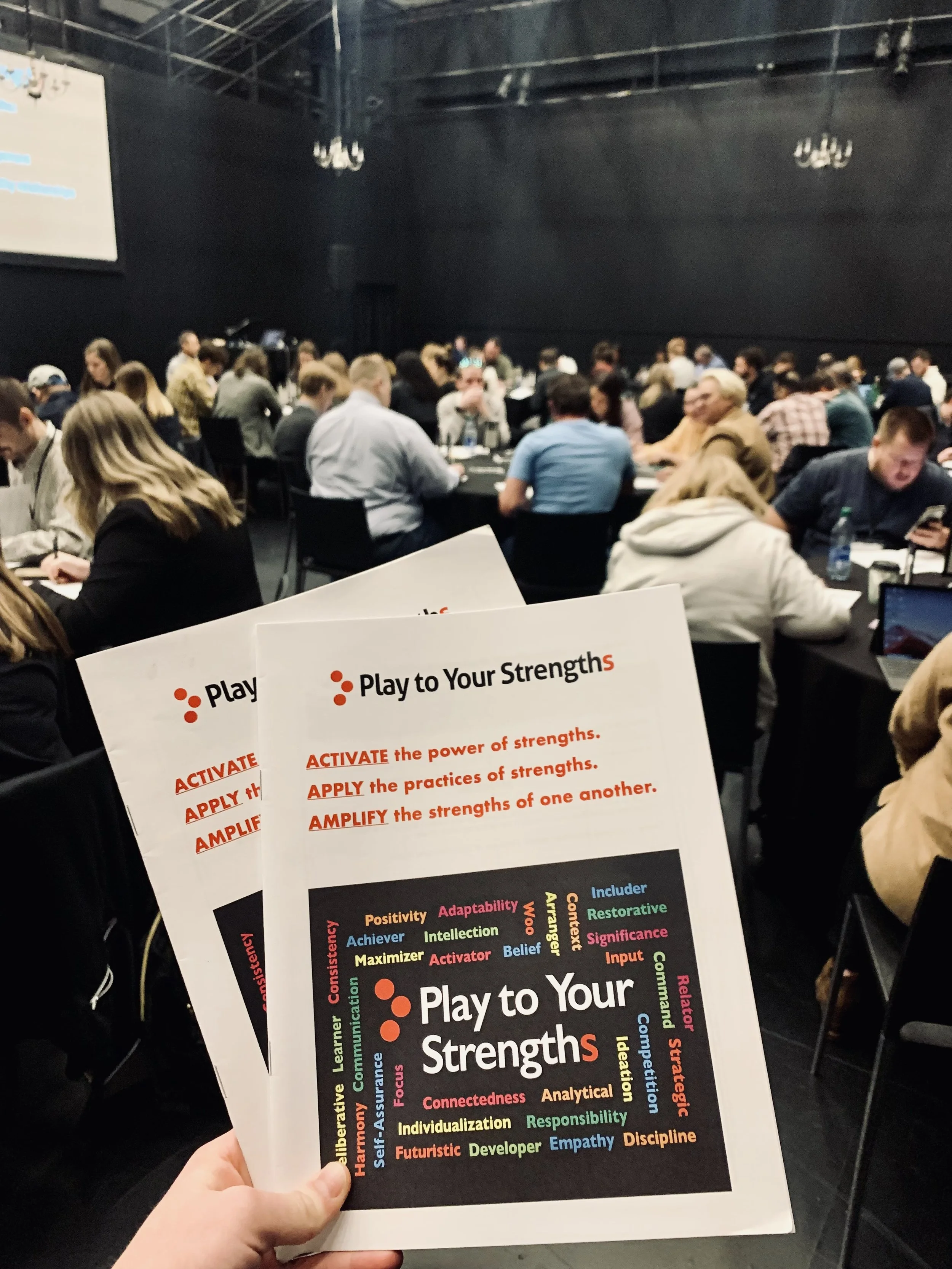 A person holding a flyer titled "Play to Your Strengths" with colorful words related to strengths such as positivity, adaptability, resilience, and creativity. In the background, there is a conference room with many people seated at tables, engaged in a group activity or workshop.