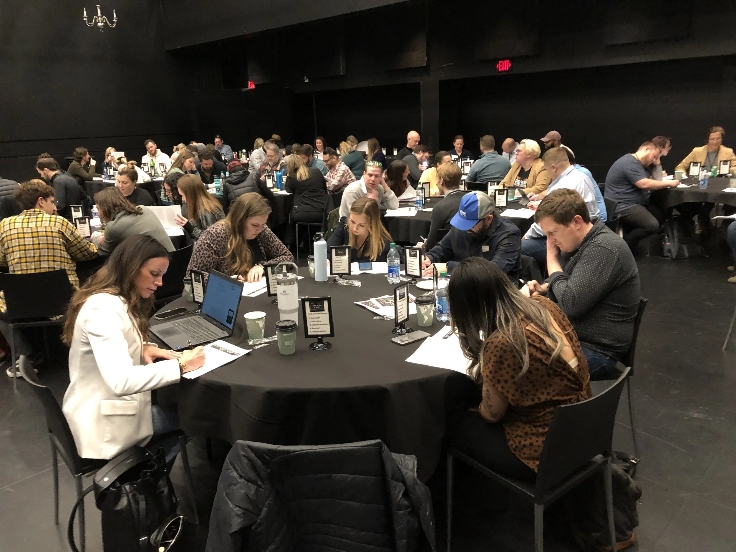 People seated around black tables in a dimly lit conference room, engaged in conversations and working on laptops and papers.