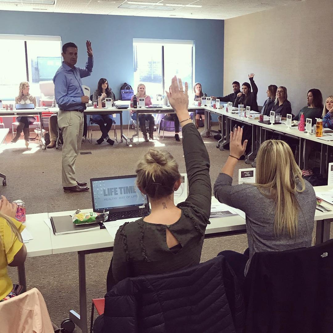 Man giving a presentation to a group of people in a conference room, with several attendees raising their hands.