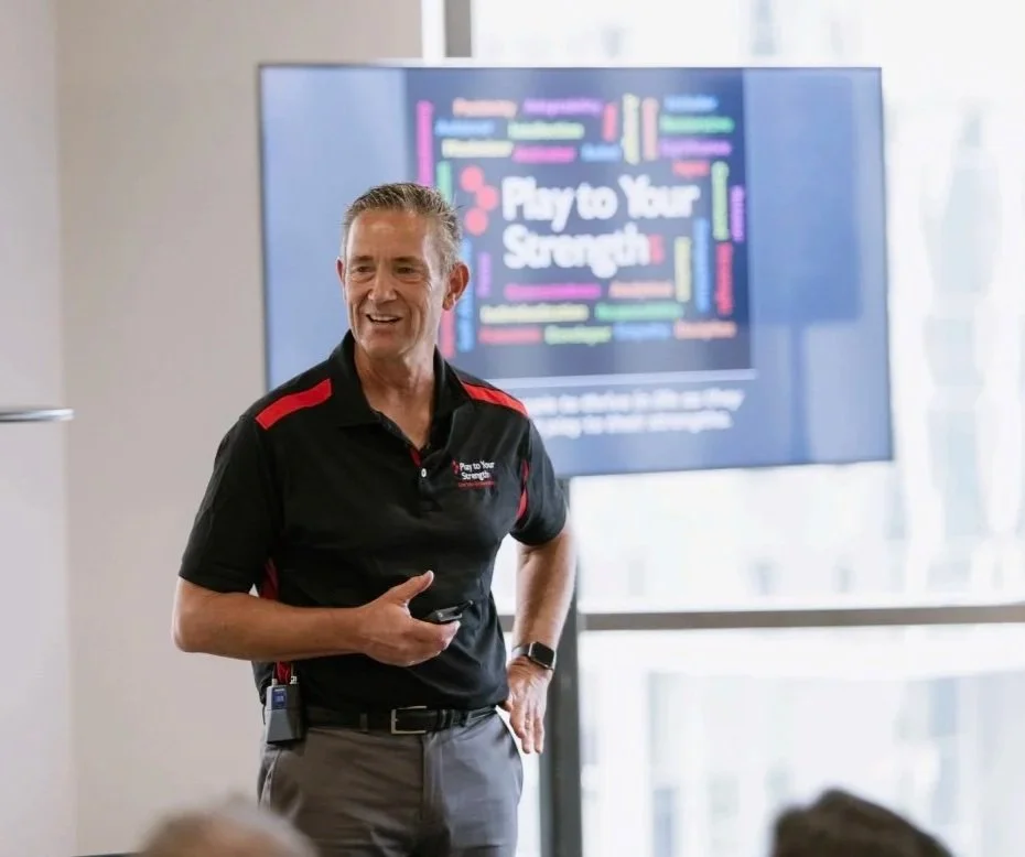 A man in a black and red polo shirt stands in front of a screen displaying a colorful word cloud, speaking to an audience.