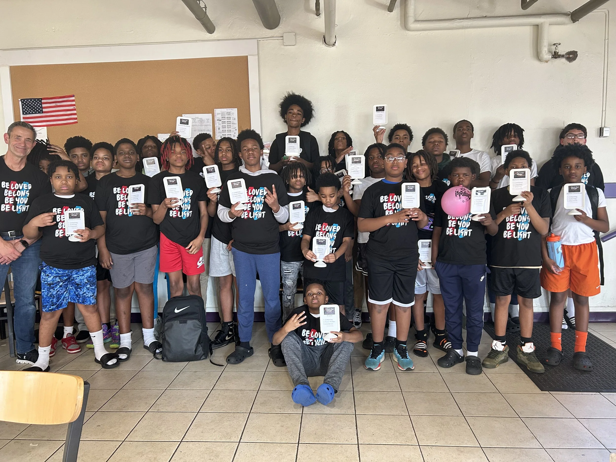 A group of children and two adults standing indoors, some holding certificates and a pink balloon. The children are wearing black T-shirts with the words "Be Loved Be You Be Light". An American flag is on the wall behind them.