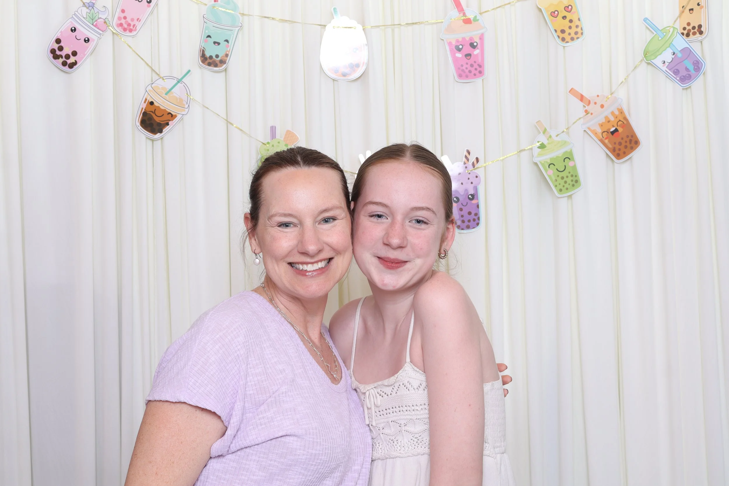 A woman and a girl smiling and posing for a photo in front of a decorated background with cartoon-style bubble tea banners hanging.