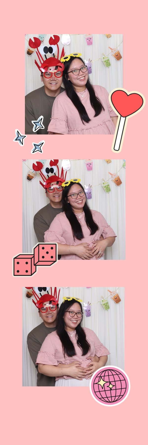 Three friends taking playful photos in a photo booth decorated with colorful paper cups, stickers, and a pink background. The man is wearing crab-themed headgear with large eyes and claws. The woman in front is wearing glasses and a pink top, smiling